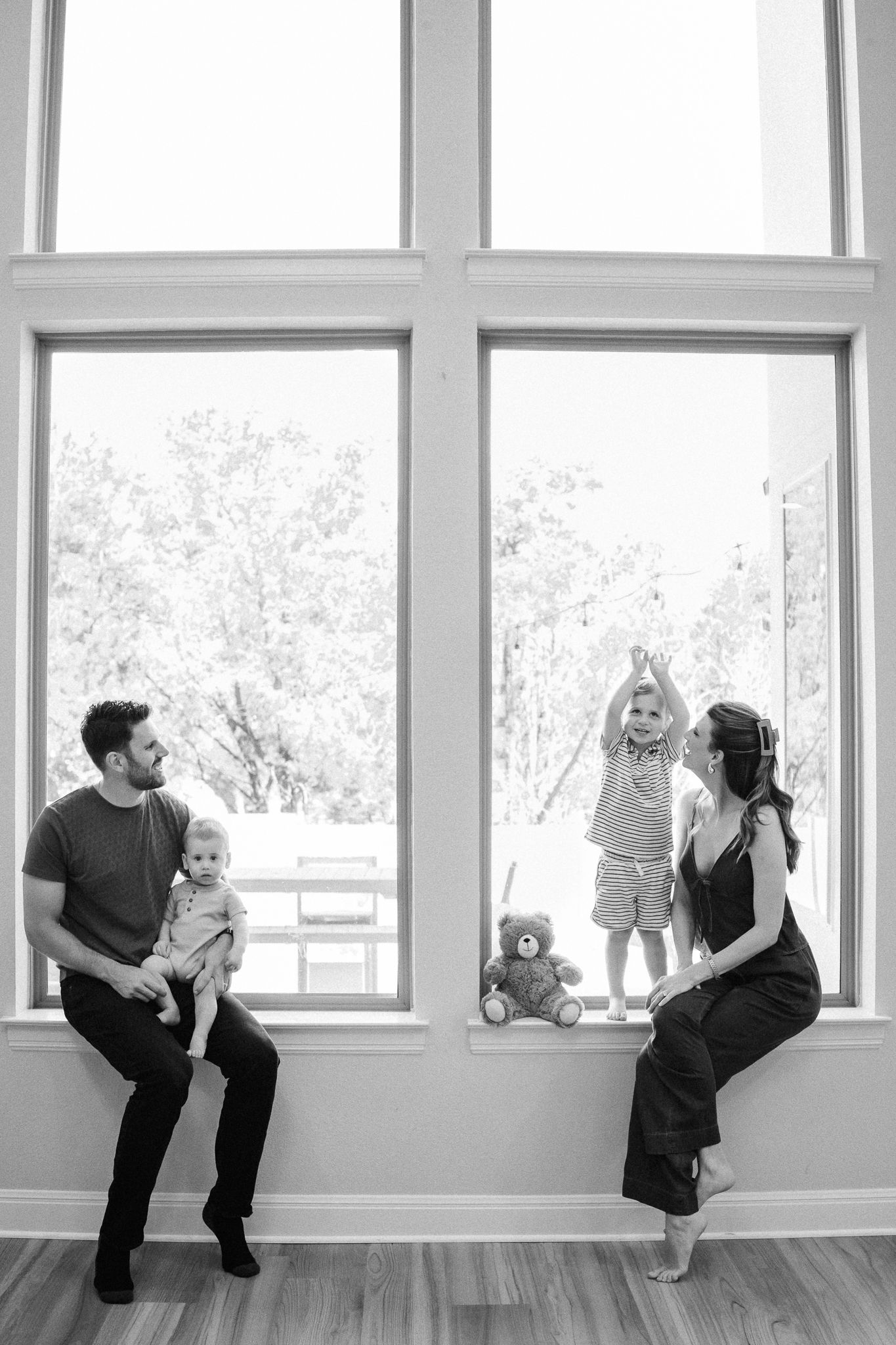 Black and white family photo with parents and two young children sitting by large windows with trees visible outside.