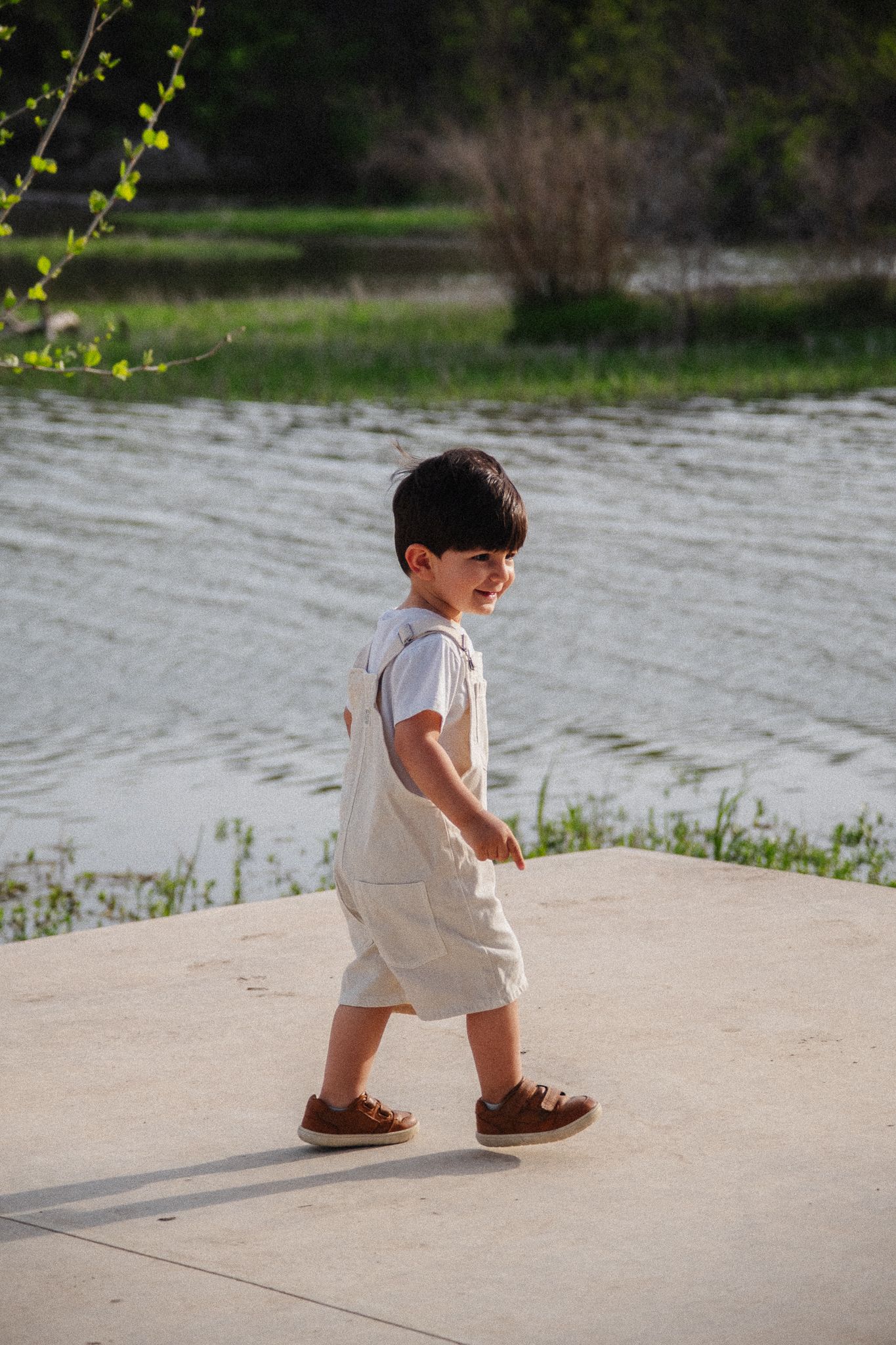 A toddler in a white outfit and brown shoes walks along a concrete path near a lakeside on a sunny day.