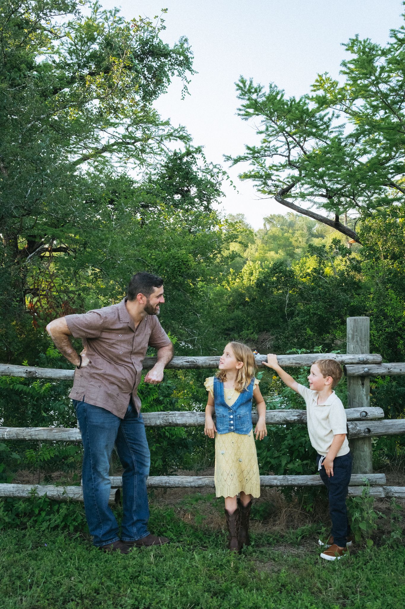A man and two children laugh together by a rustic wooden fence surrounded by lush green trees outdoors.