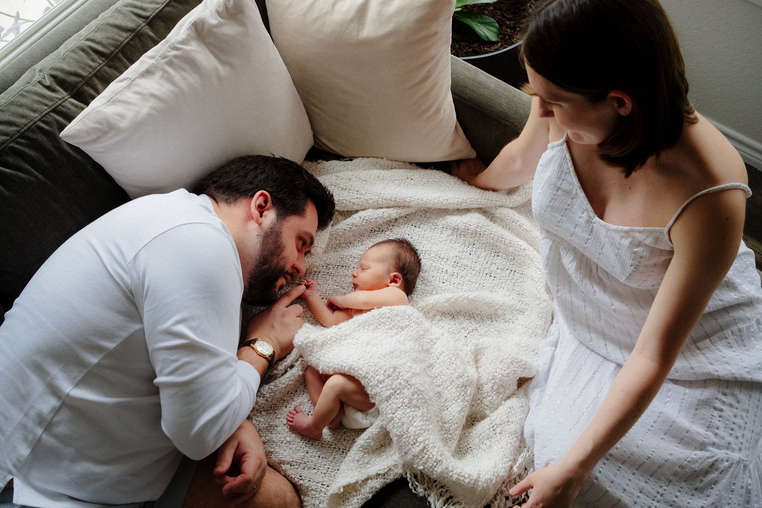 New parents lovingly gaze at their newborn baby lying on a blanket between them on a couch.