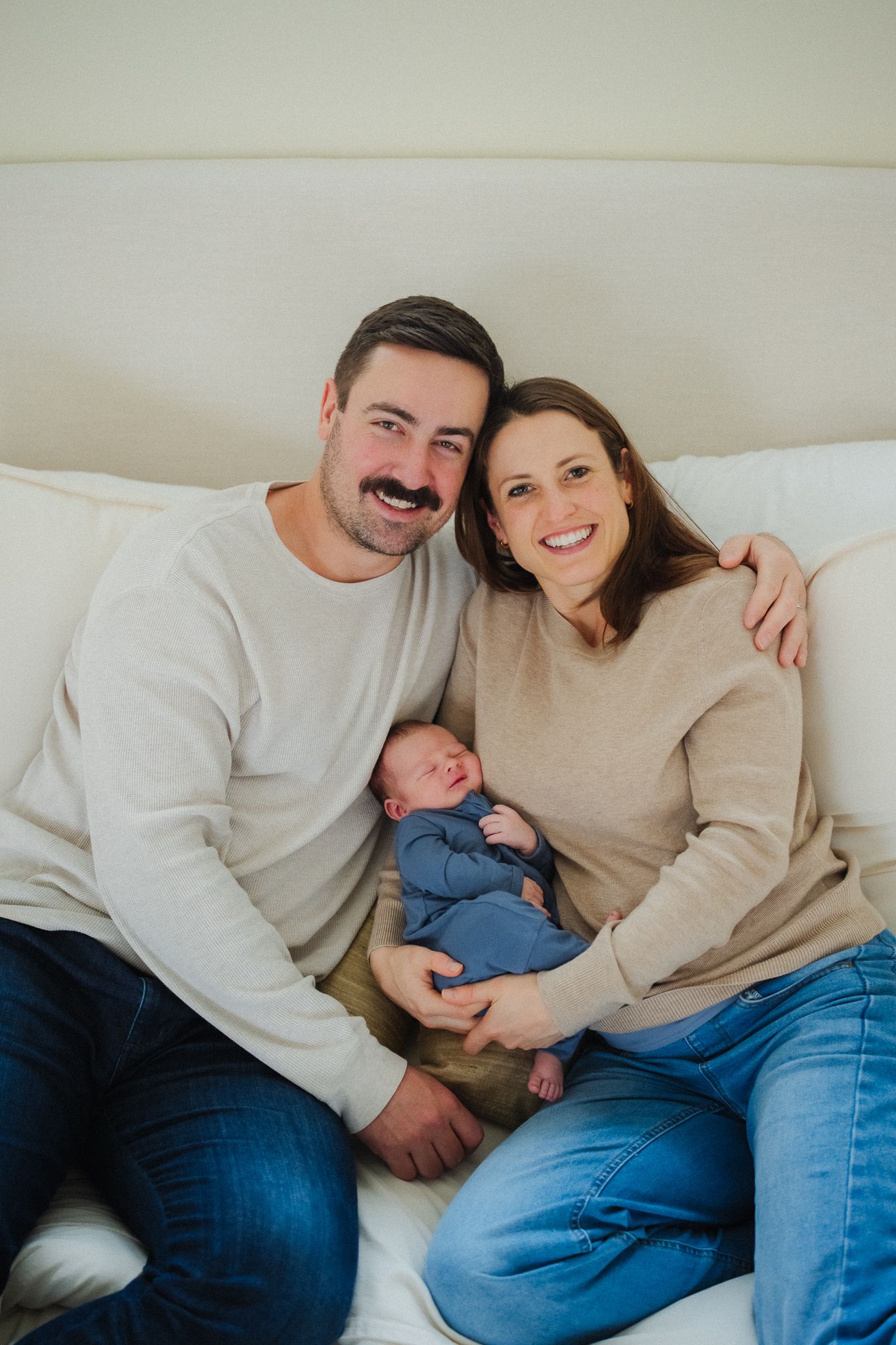 Happy couple sitting on bed smiling, holding their newborn baby dressed in blue.