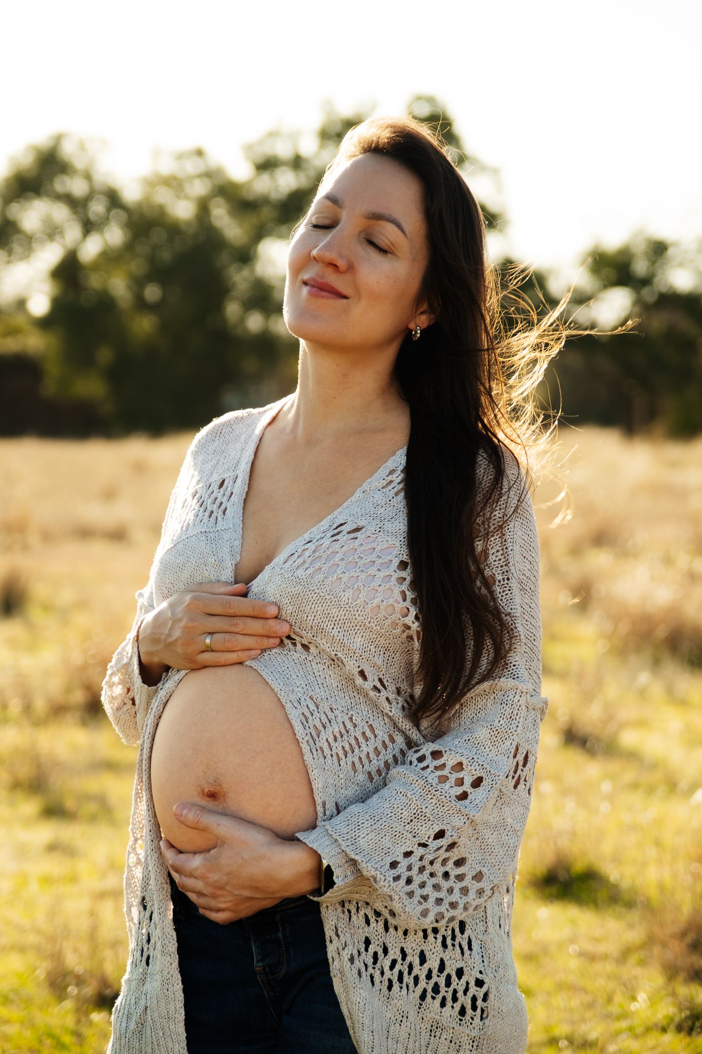 Pregnant woman in white crochet cardigan standing in golden field at sunset with hands on belly.