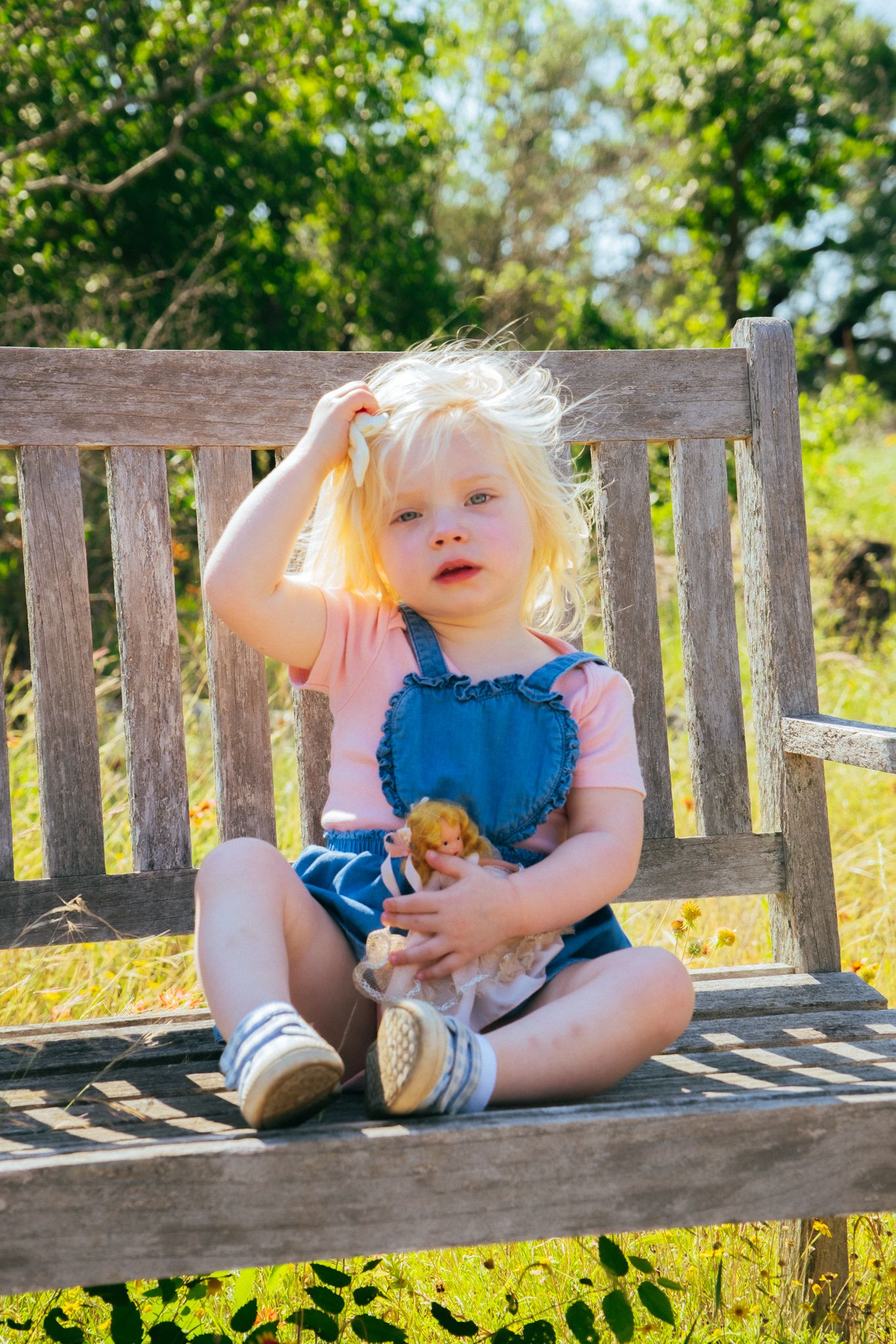 A young child in overalls and pink shirt relaxes on a wooden park bench during a sunny afternoon.