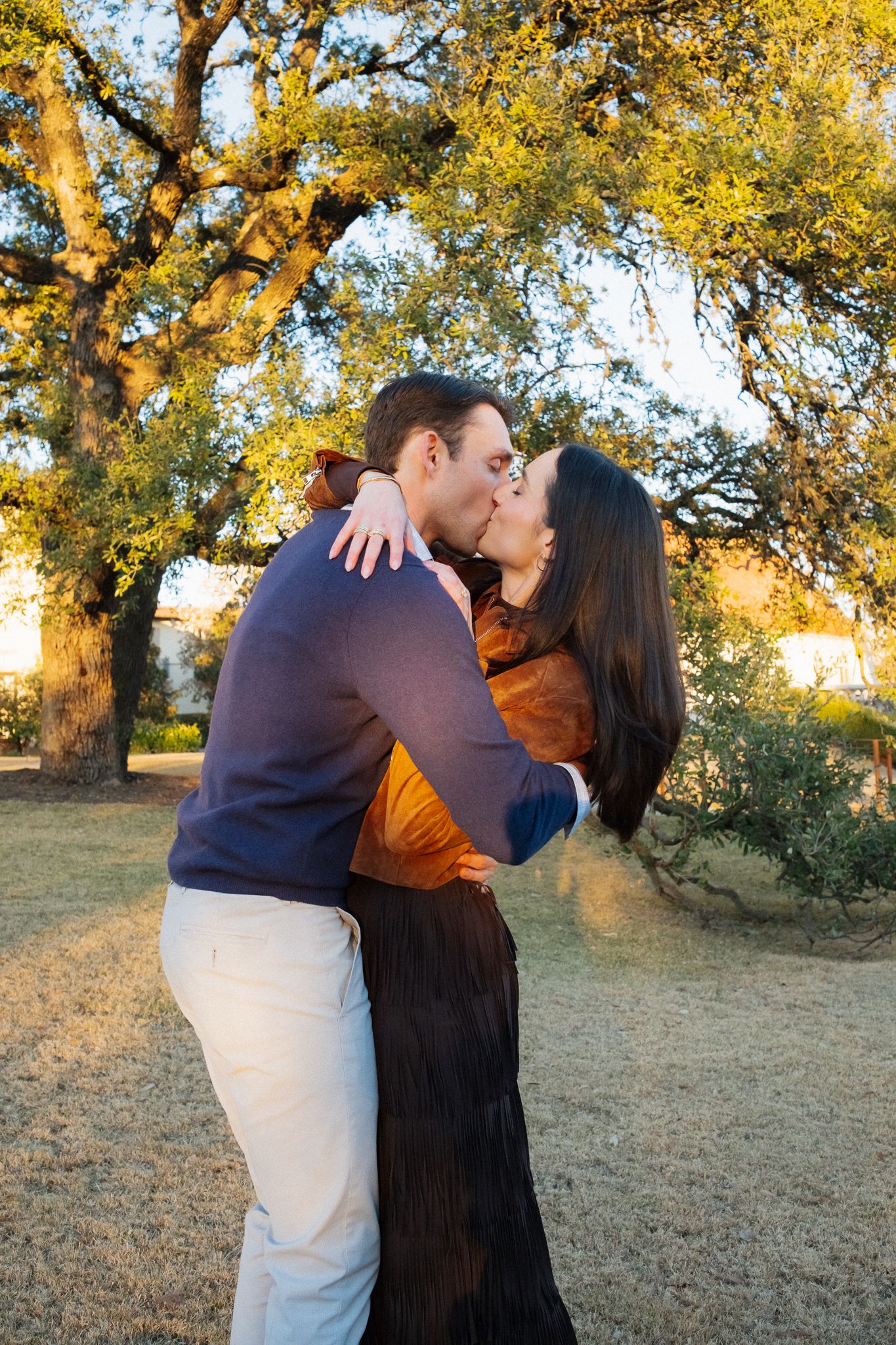 Couple sharing a romantic kiss outdoors beneath large oak trees in golden hour sunlight, woman in orange top and black skirt.