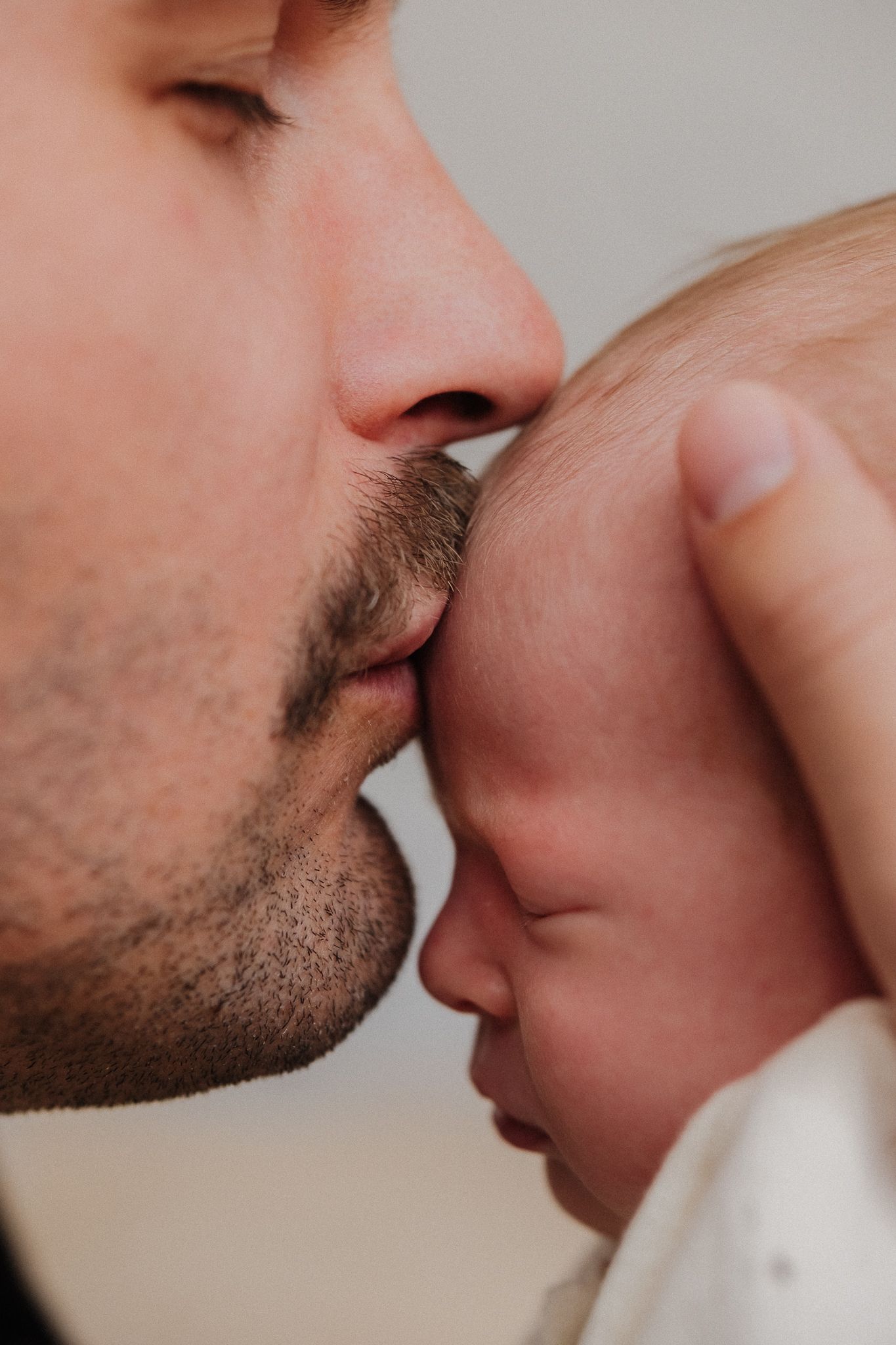 Close-up of a gentle forehead kiss being given to a newborn baby wearing white clothing.