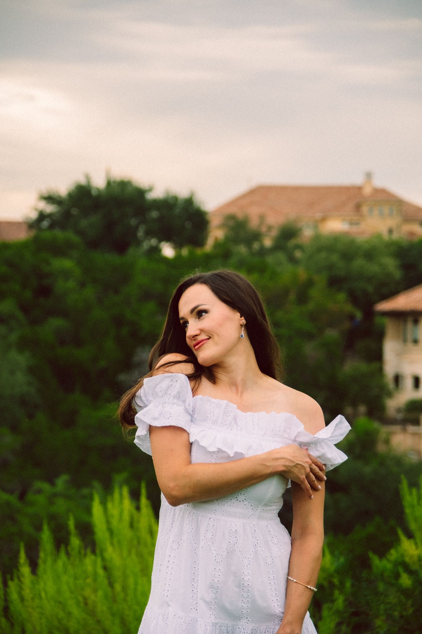 A person in a white ruffled dress poses outdoors against a backdrop of green trees and residential buildings.