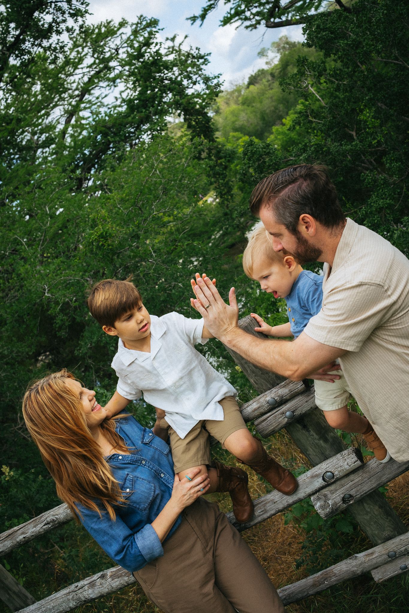 Family of four enjoying outdoor time near a wooden fence surrounded by lush green trees on a sunny day.