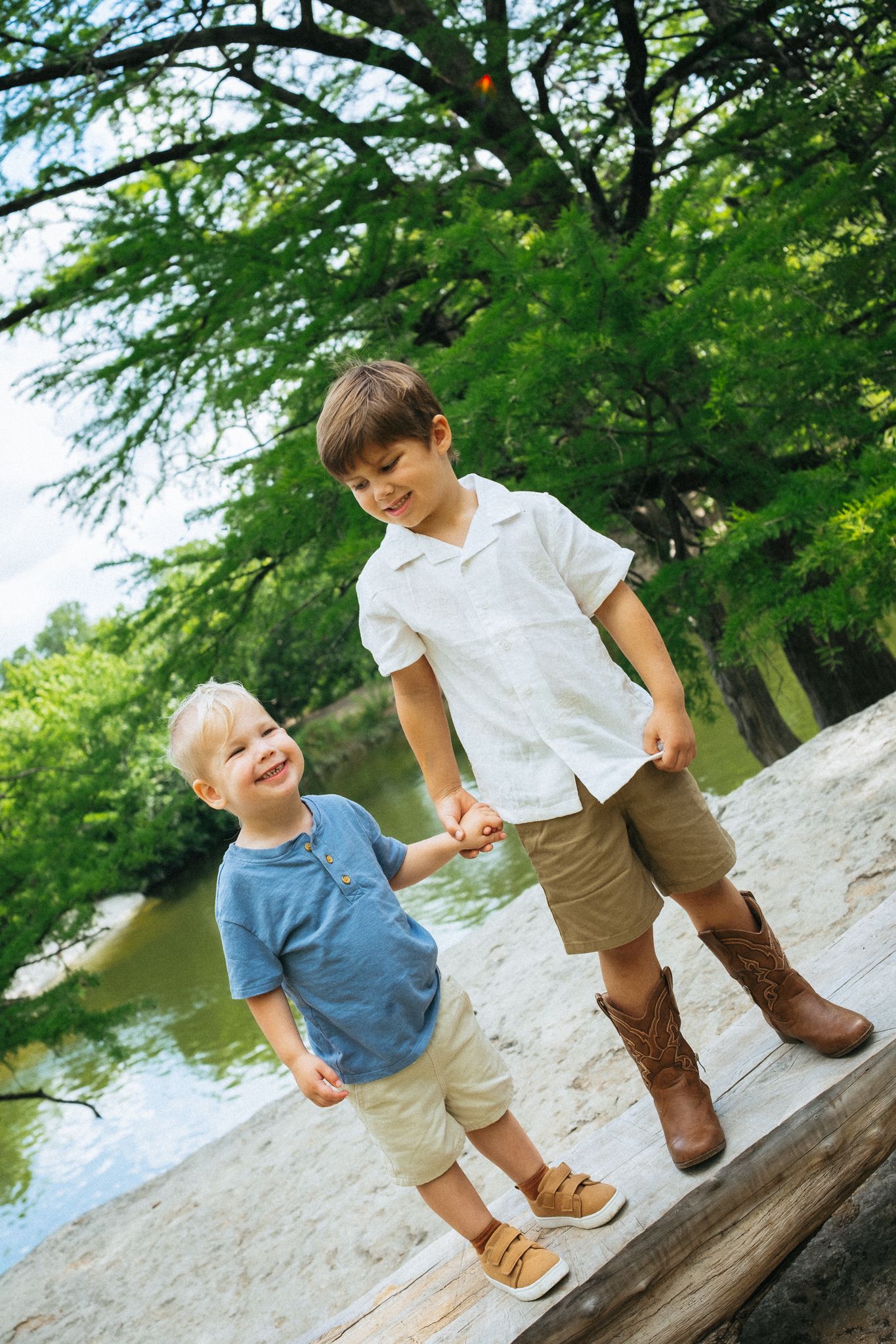 Two young boys holding hands outdoors near a river, smiling, wearing casual summer clothes among green trees.