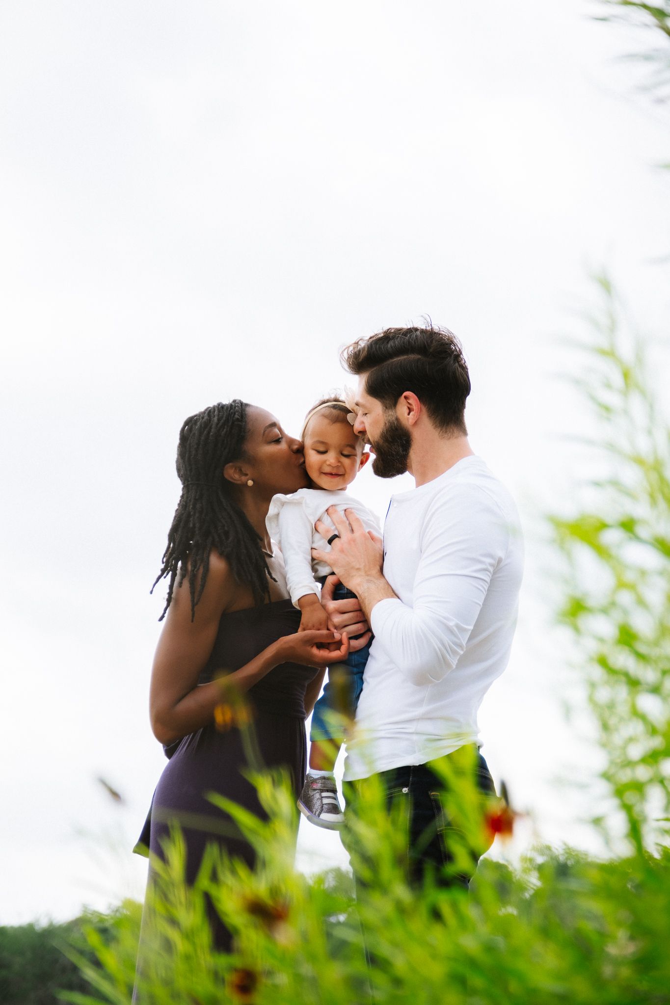 A couple stands together in a meadow holding their baby while green plants sway in the foreground.