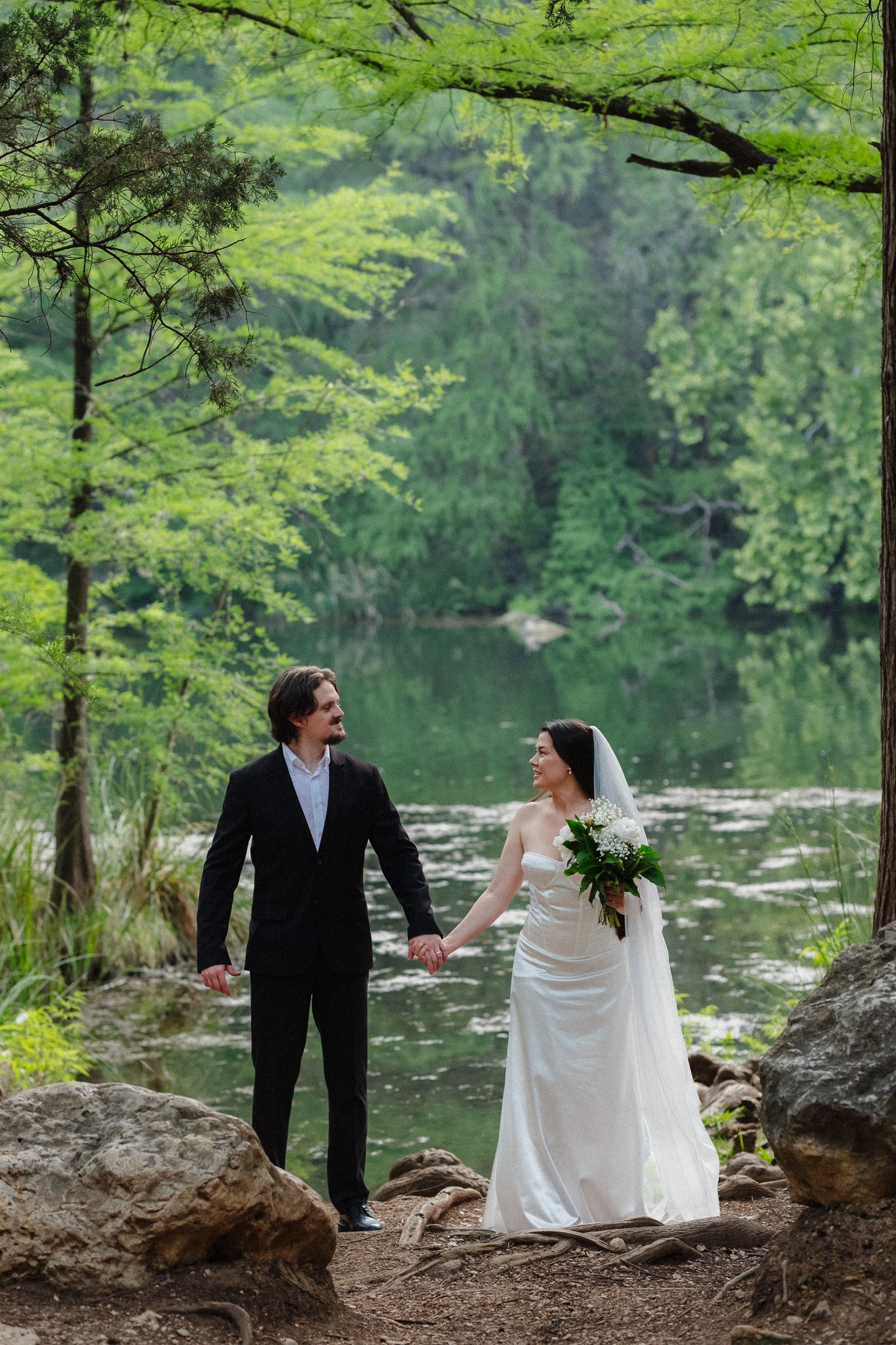 Bride and groom hold hands by a serene forest pond, she in white gown with veil, he in black suit.