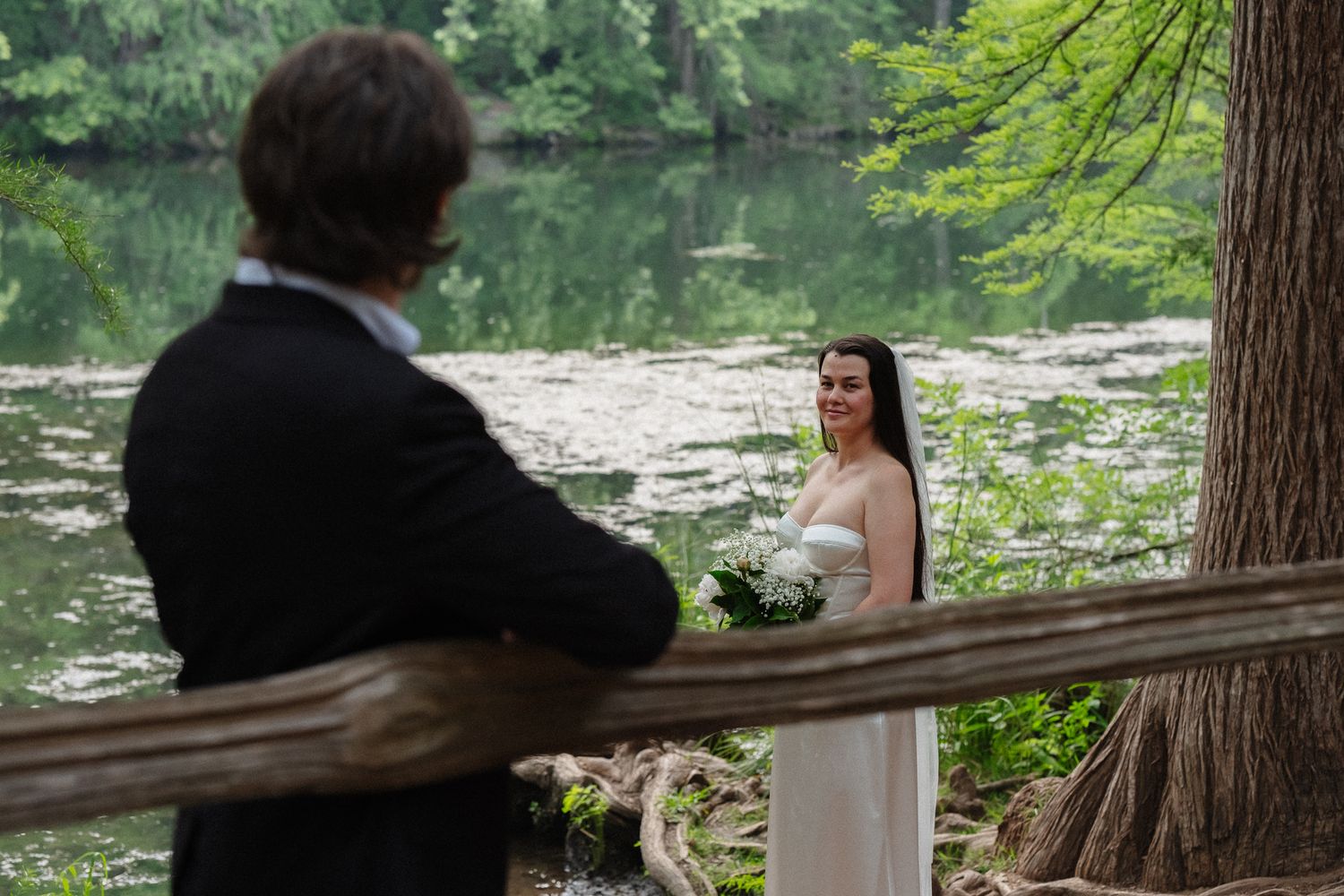 Bride in white dress holds bouquet near a serene pond in a lush green forest during an outdoor wedding ceremony.