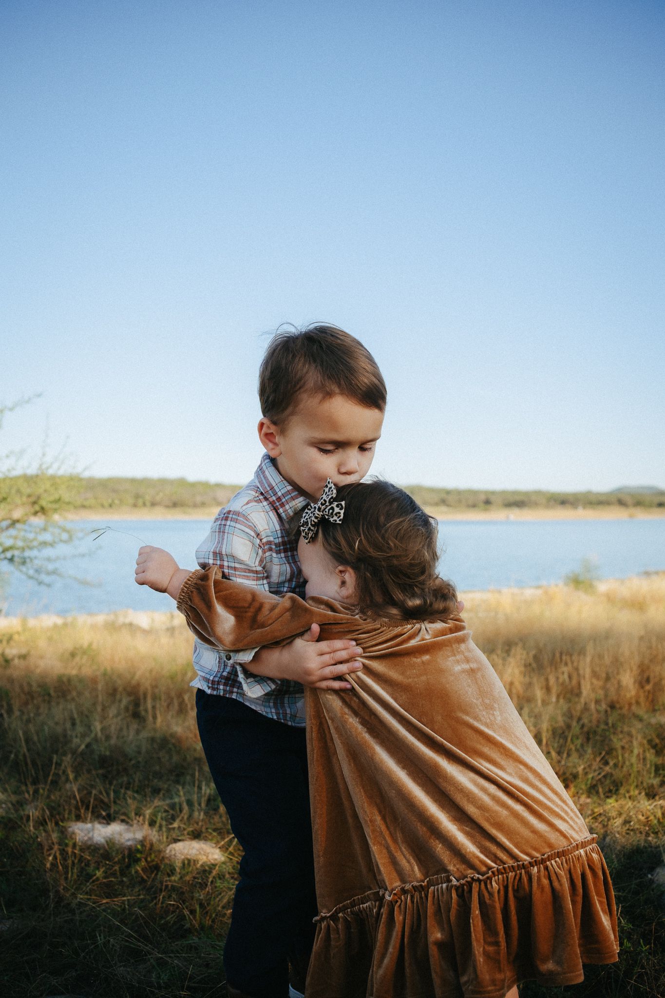 A series of photos showing a young child hugging someone in a brown dress in a sunlit outdoor field by a lake.
