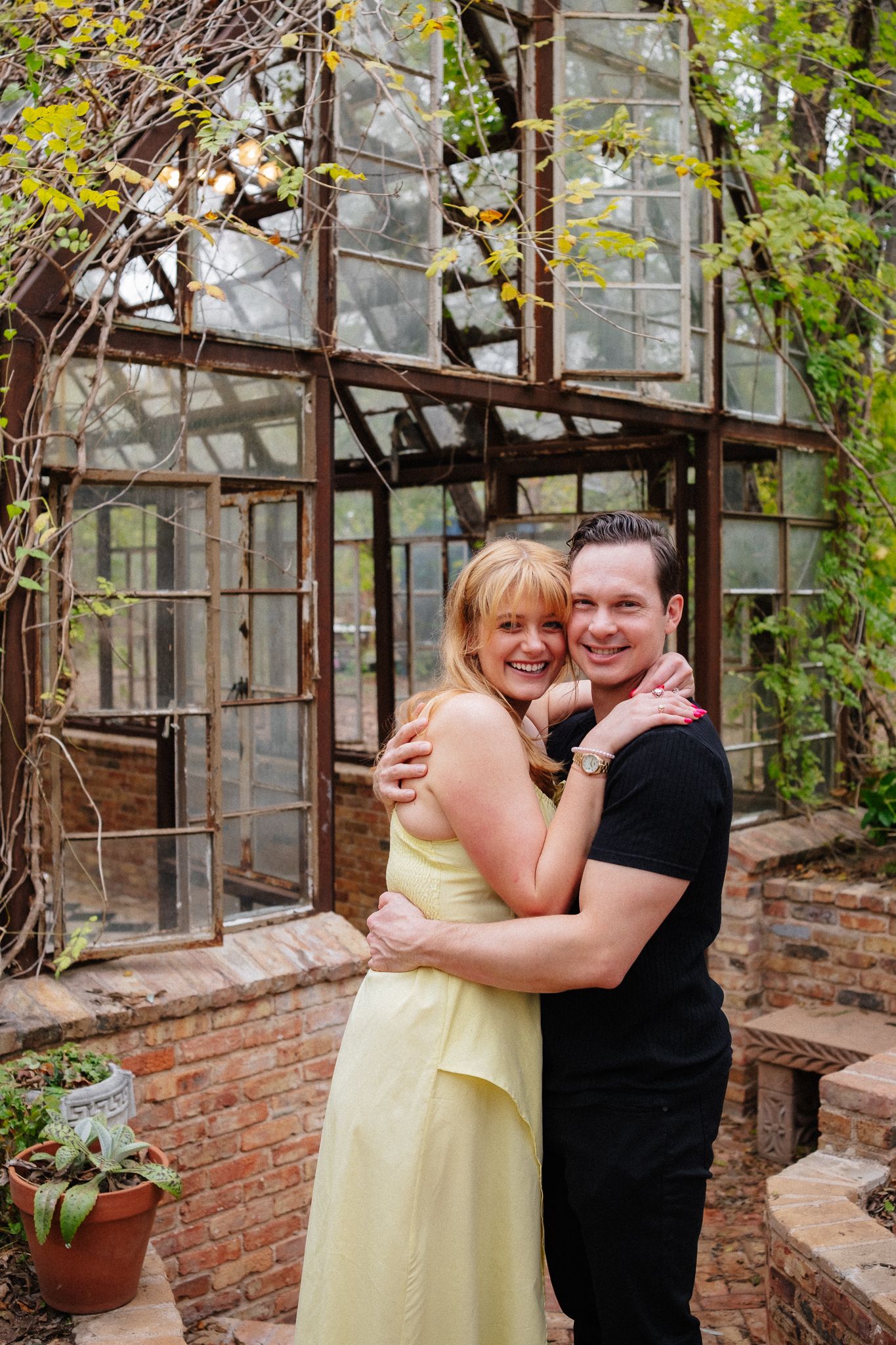 A couple embraces in a rustic greenhouse with brick walls and lush greenery surrounding them.