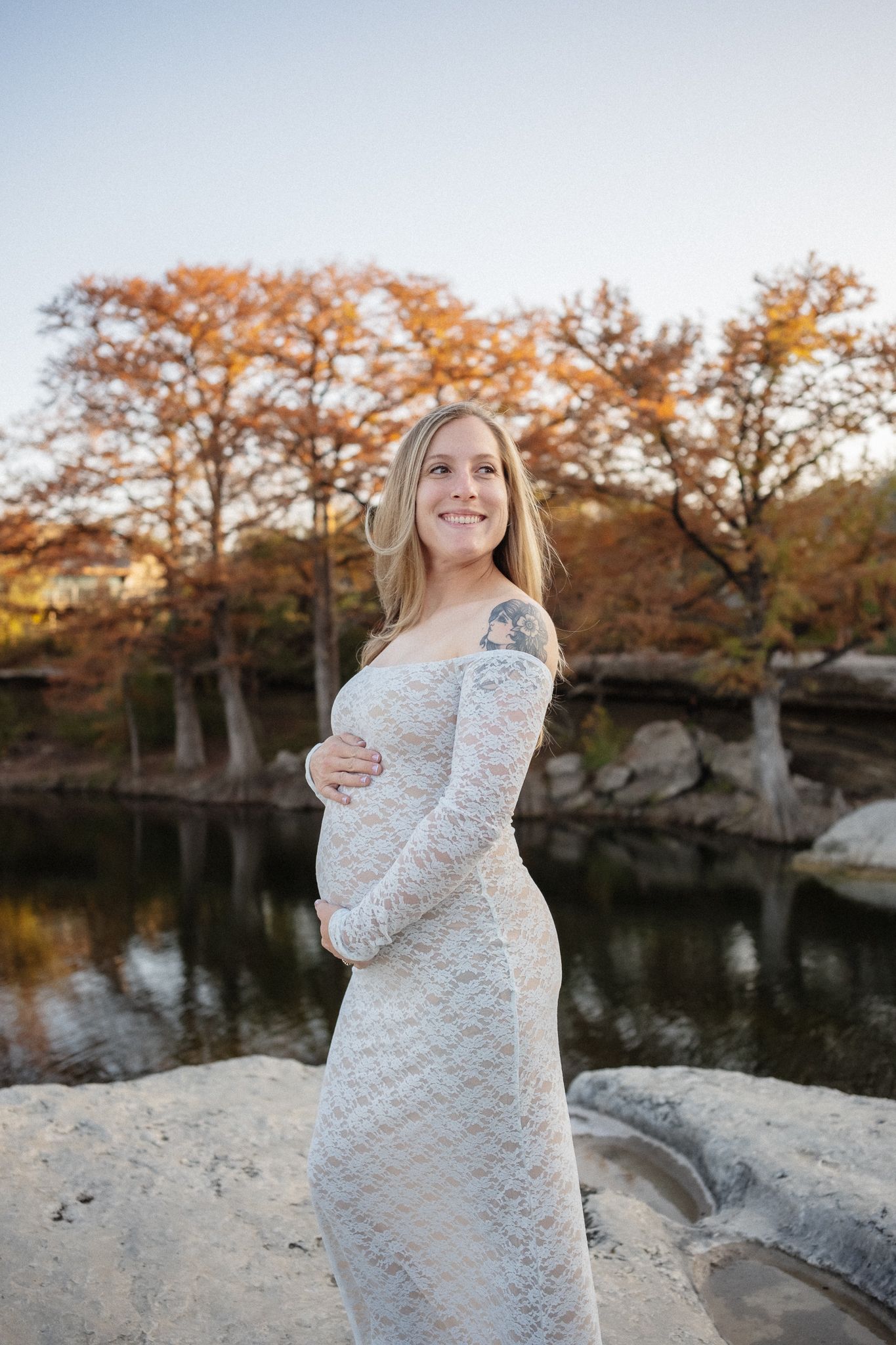 A pregnant woman in a white lace dress poses by a pond during autumn with orange trees in the background.