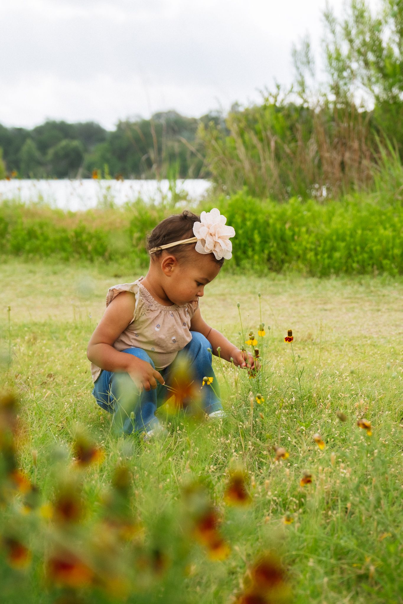 A child in blue clothing crouches down to examine wildflowers blooming in a sunny meadow.
