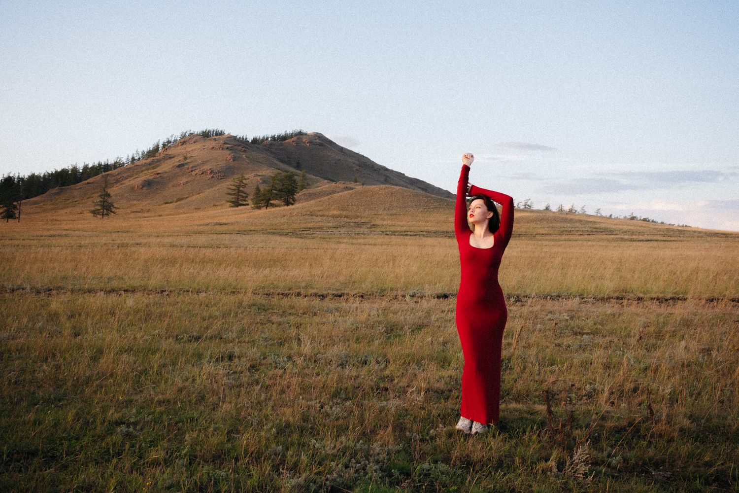 Woman in red dress poses gracefully in open field with rolling hills and clear sky backdrop.