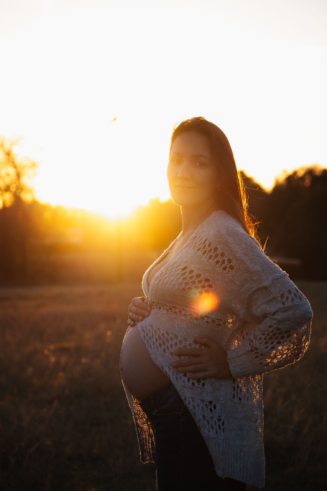 Silhouette of pregnant woman in crochet sweater stands backlit by warm golden sunset light outdoors.