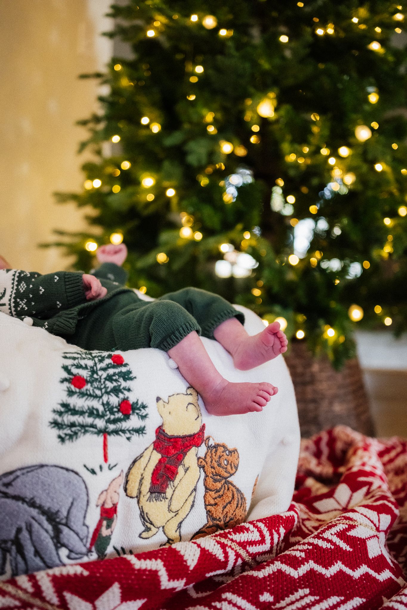 Baby feet resting on a Winnie the Pooh pillow near a lit Christmas tree with red holiday blanket.
