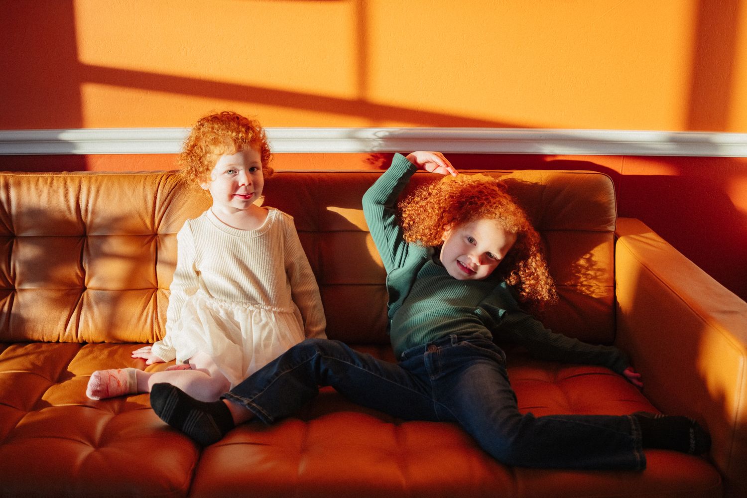 Two red-haired children laughing and playing on a brown leather sofa in warm sunlight.