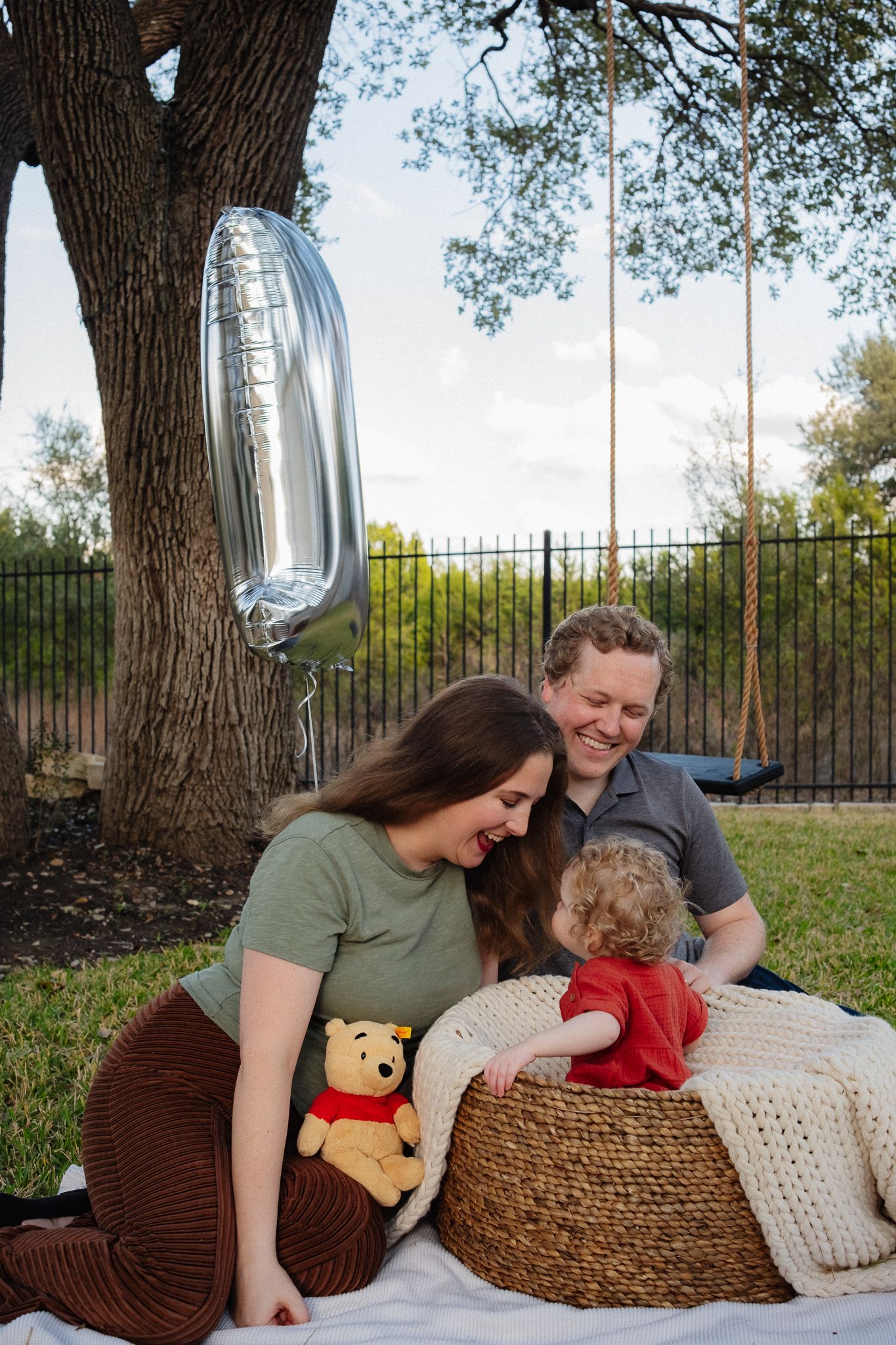 Family relaxing together on blankets with a Winnie the Pooh plush toy under a shady tree.