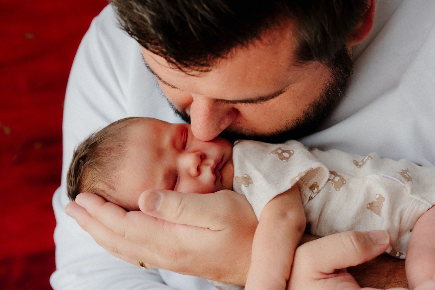 Bearded father tenderly kissing his sleeping newborn baby cradled in his hands against a red background.