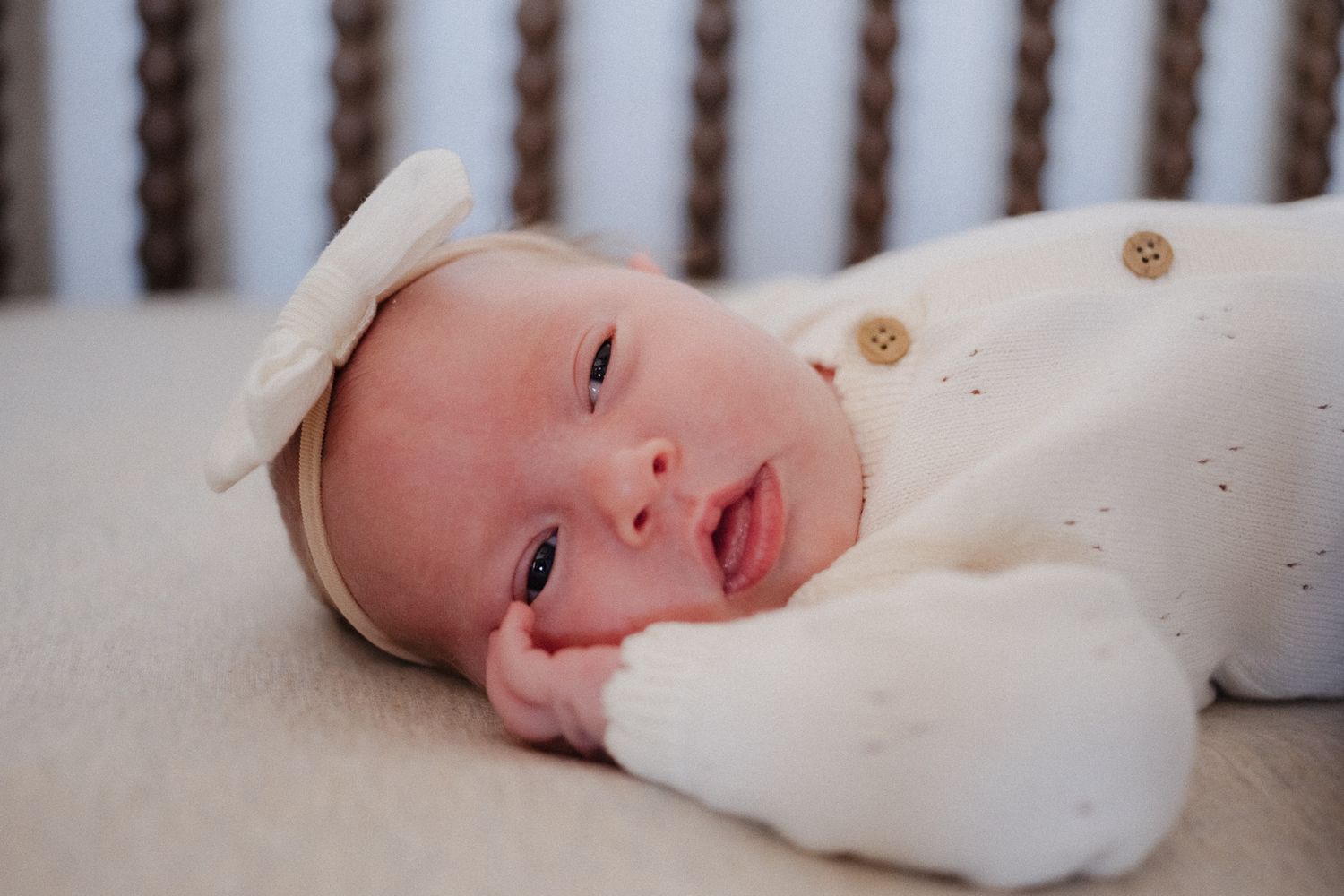 Newborn baby in white knit outfit and bow headband lying in a crib, smiling softly.