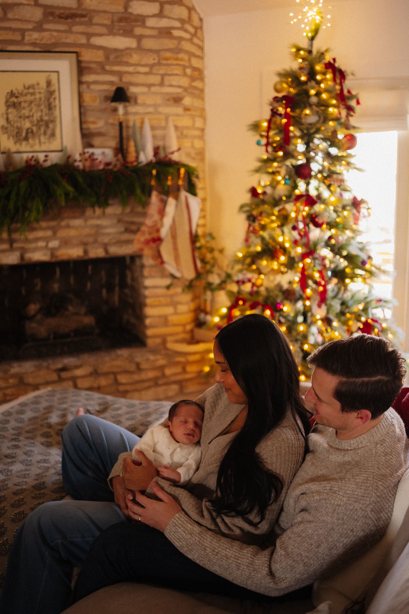 Young couple sitting on couch holding newborn baby in front of decorated Christmas tree and stone fireplace.