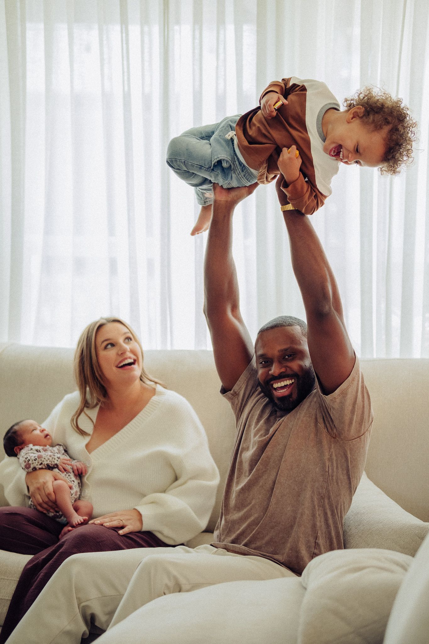 Happy interracial family on white sofa, dad lifting toddler overhead while mom holds newborn baby, laughing together at home.