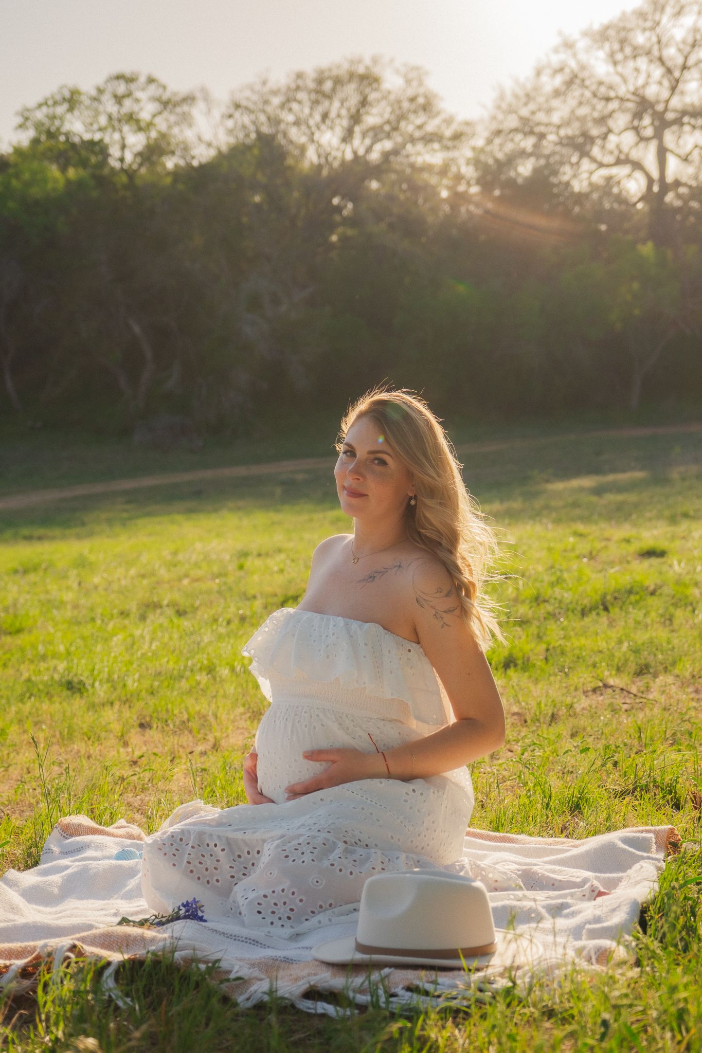 Pregnant woman in white eyelet dress sitting on blanket in sunlit green field, cradling baby bump during outdoor maternity photo.