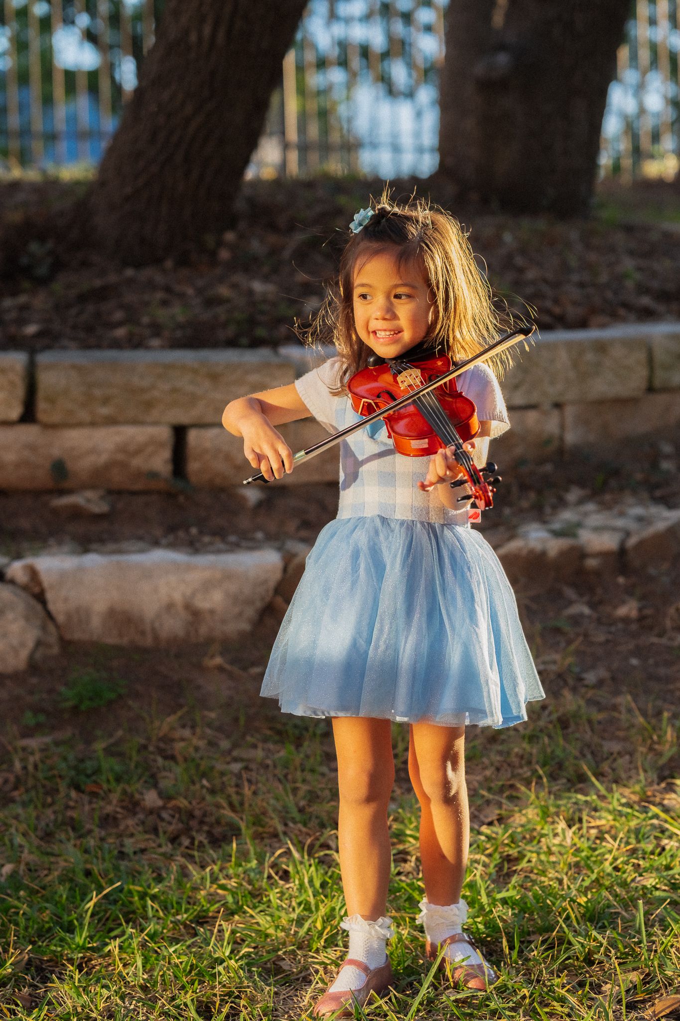 Young child playing violin outdoors in a blue dress against a natural backdrop.