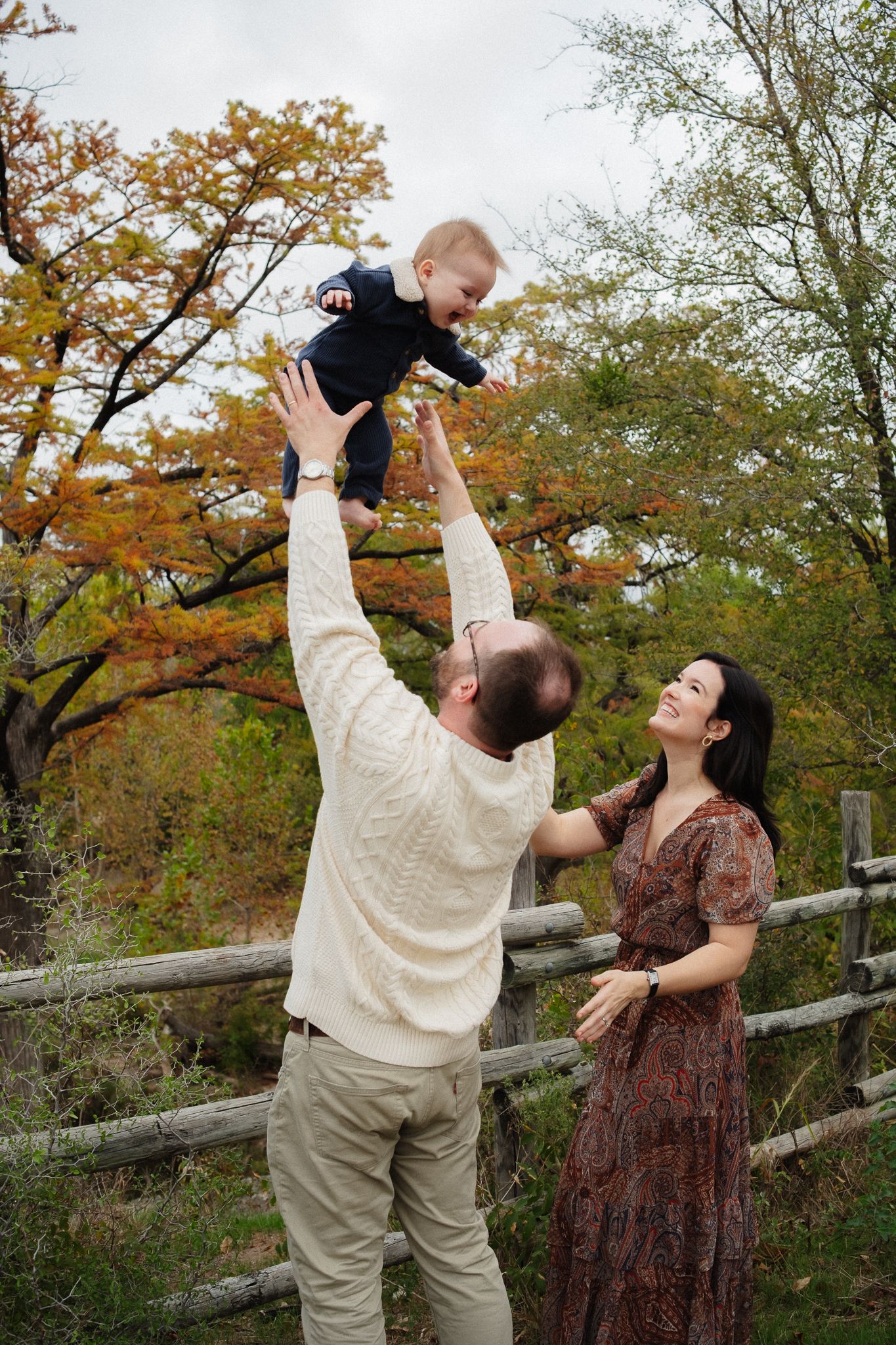 A family plays and laughs together near a wooden fence surrounded by autumn trees during an outdoor photo session.