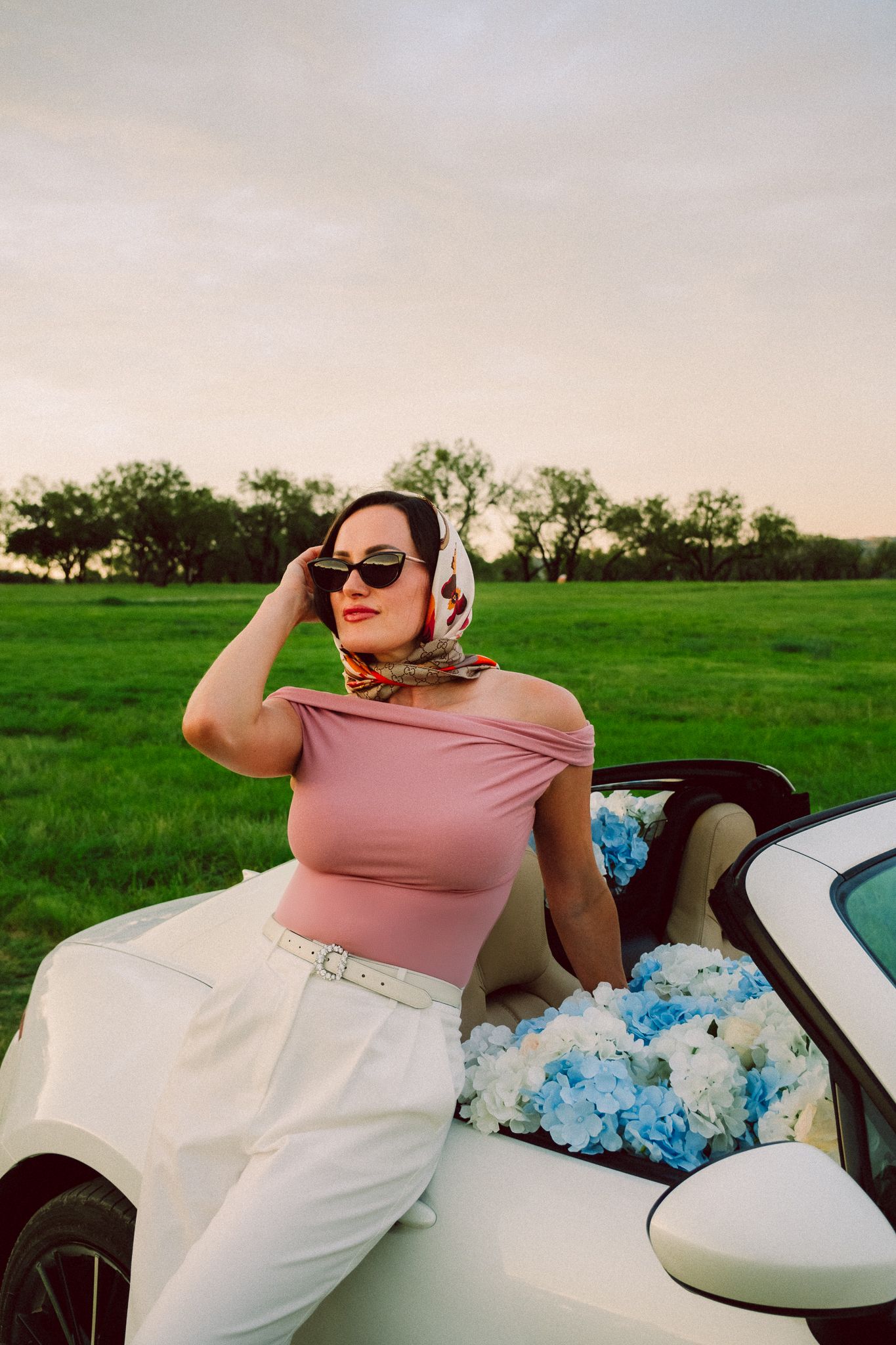 A person in sunglasses and pink top reclines against blue hydrangeas in a white convertible car on a sunny day.