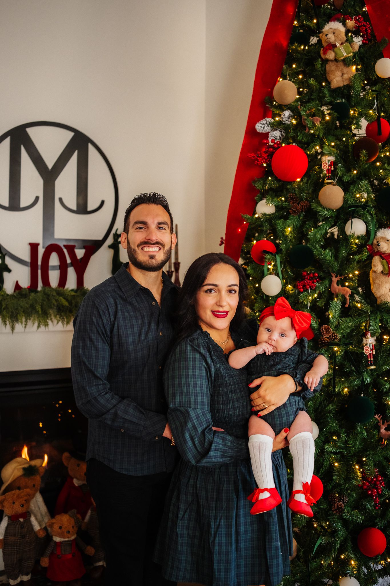 Family poses together in front of a decorated Christmas tree with baby wearing festive red outfit.