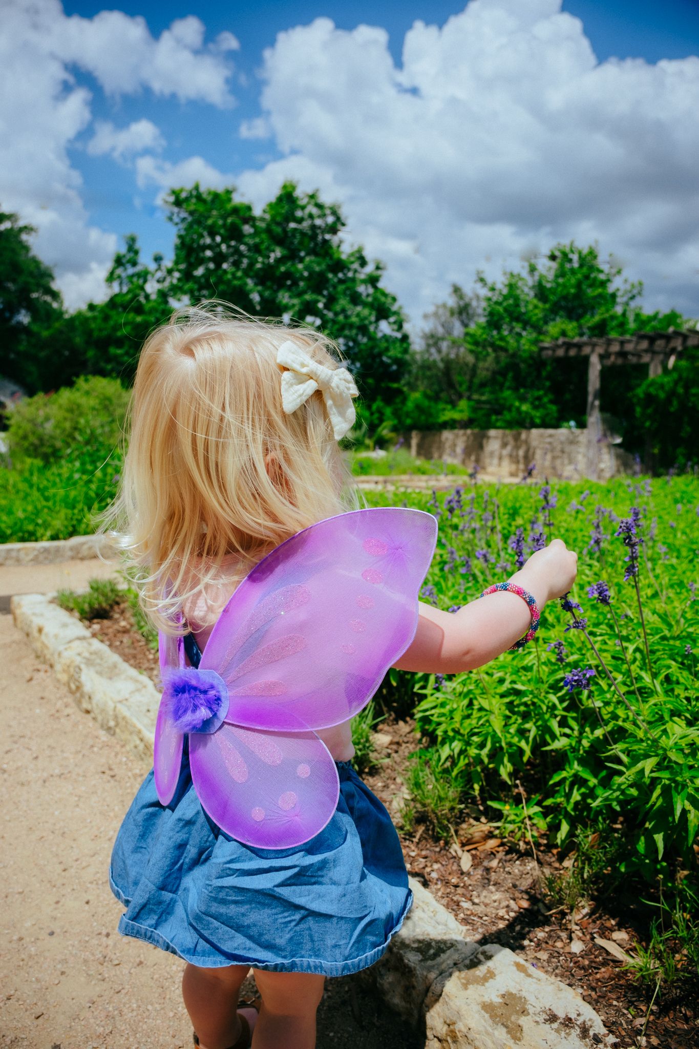 A small child wearing pink fairy wings and denim dress explores a garden path on a sunny day with fluffy clouds overhead.