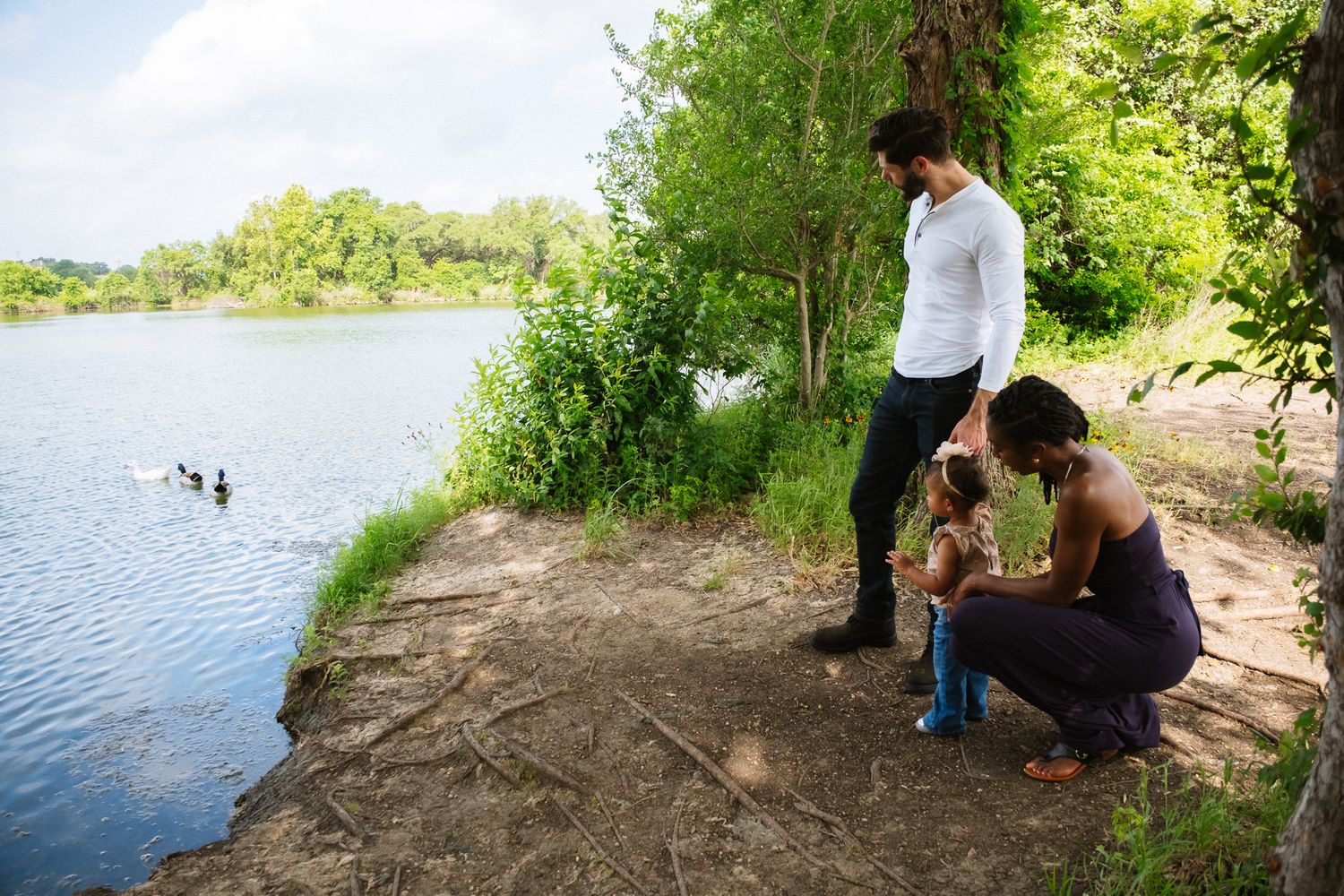 Two people in casual attire feed ducks at the edge of a scenic lake on a sunny summer day.