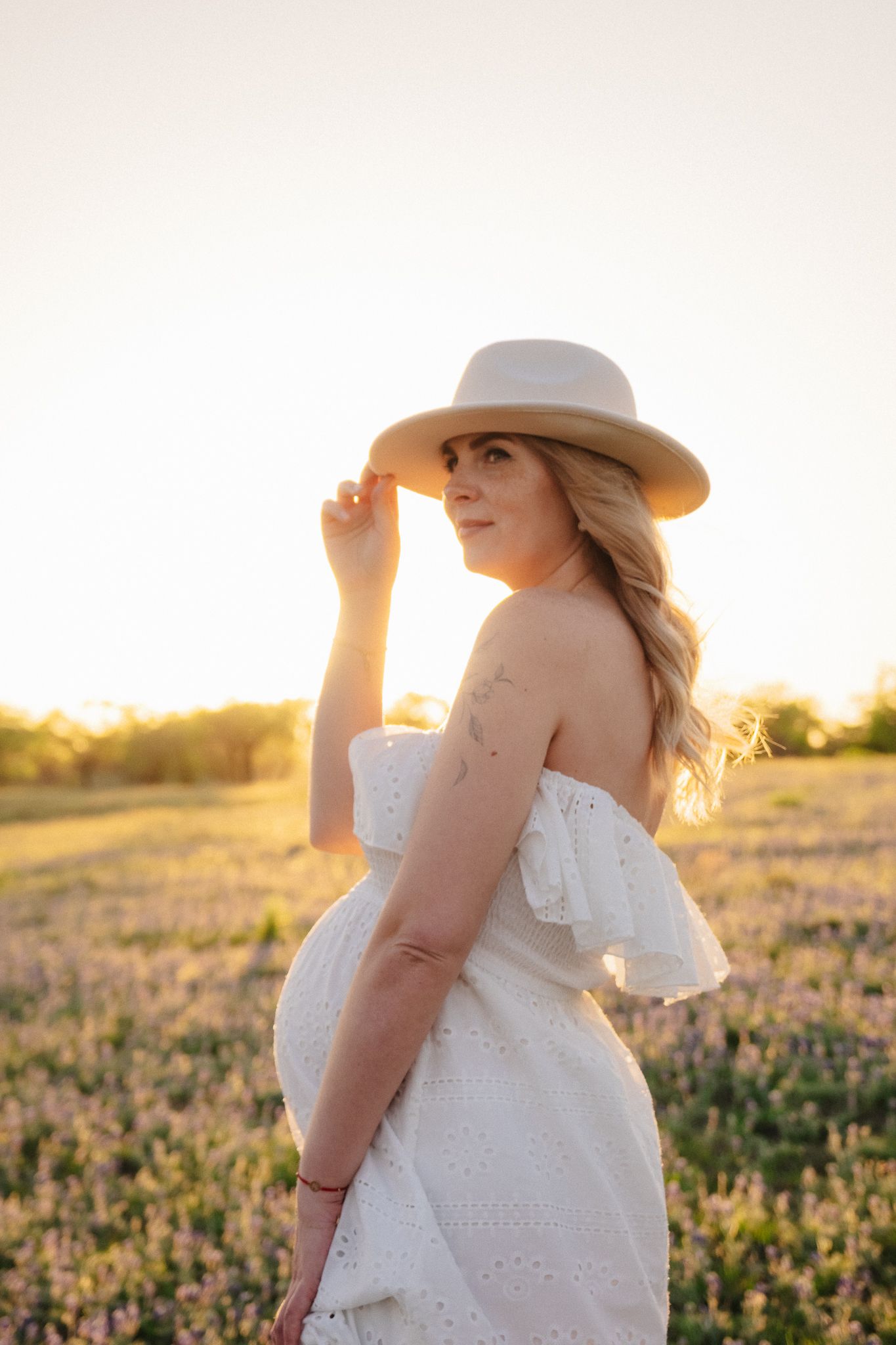 Someone in a white eyelet dress and gray hat poses in a golden sunlit field at sunset.