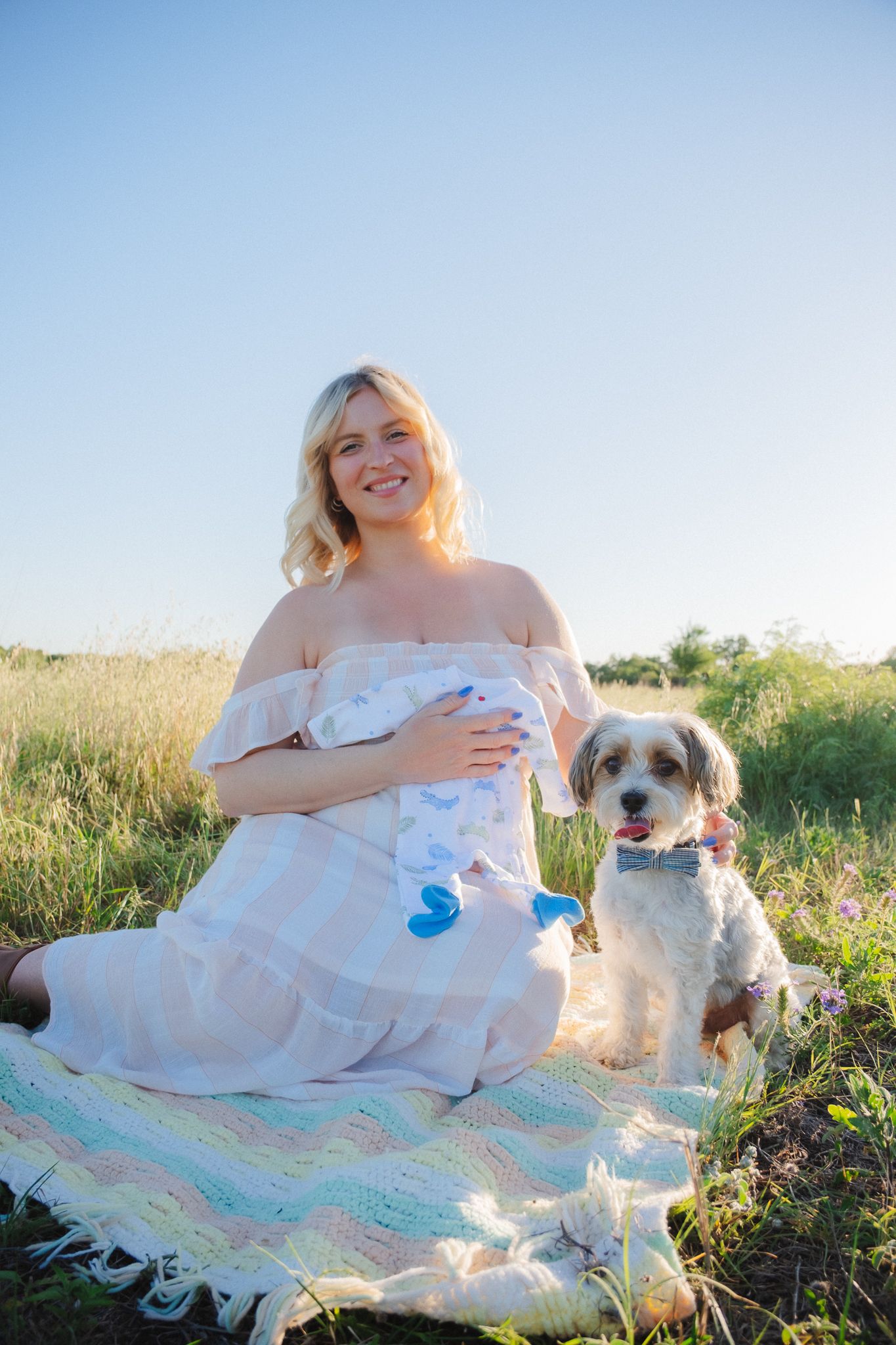Person in white dress sits on blanket in sunny field with small brown and white dog companion.