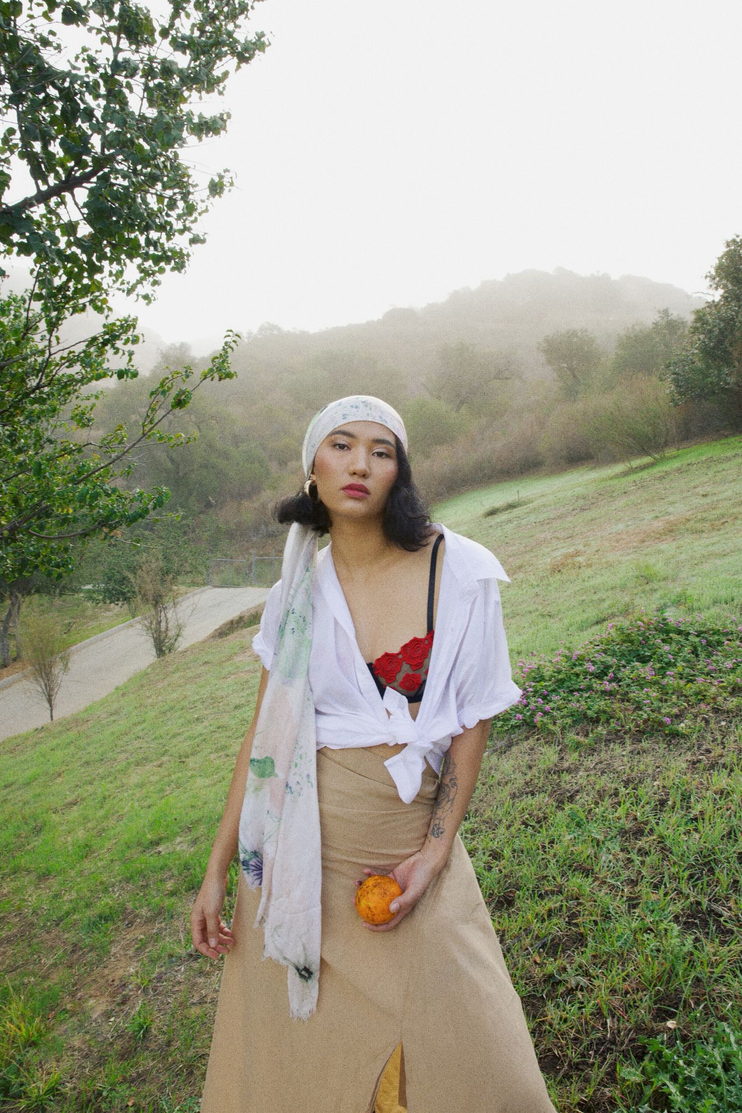 A figure poses on a misty hillside wearing a white headscarf, tan skirt, red top and light shirt in an outdoor setting.