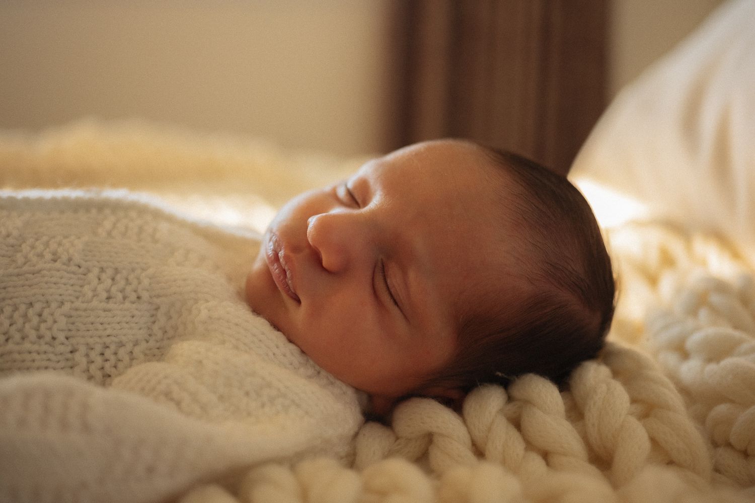 Peaceful newborn baby sleeping soundly wrapped in a soft cream chunky knit blanket in warm natural light.