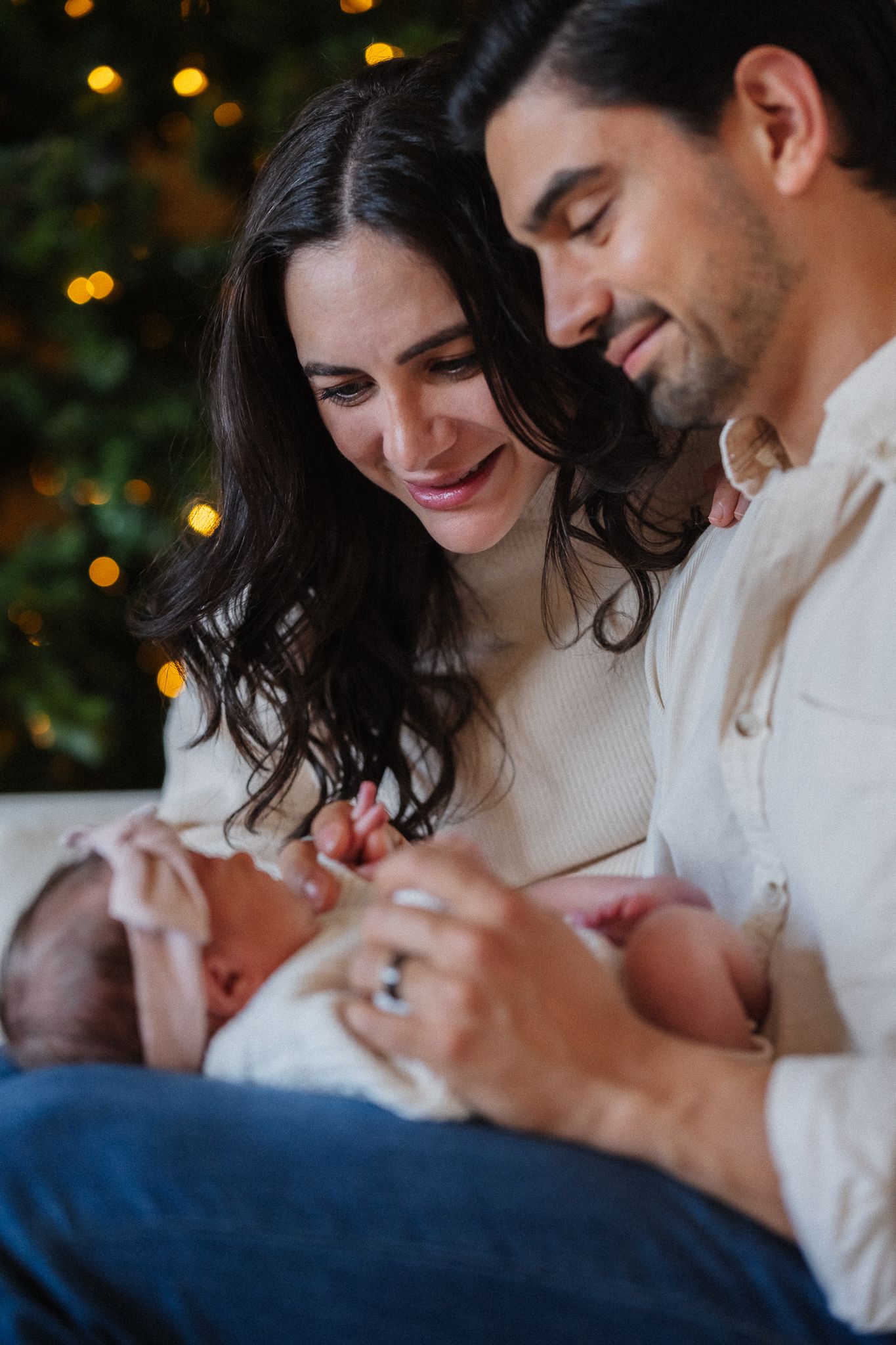 Happy couple smiling at their newborn baby near a Christmas tree with warm bokeh lights.