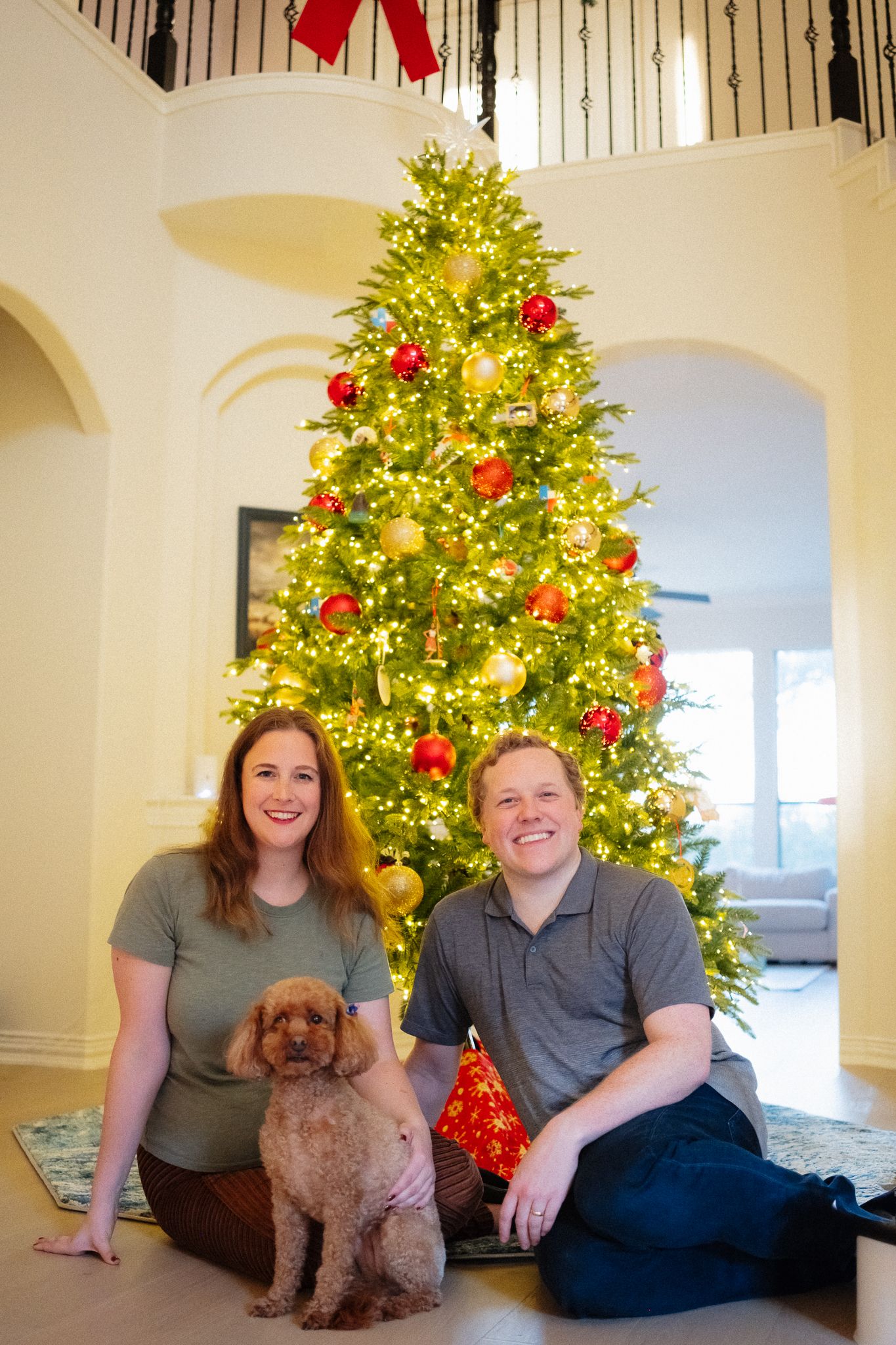 Couple sitting with brown poodle in front of decorated Christmas tree with red and gold ornaments indoors.