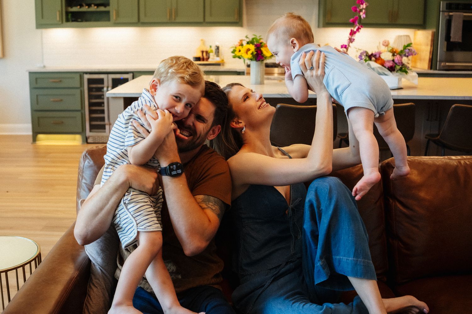 Happy family with two young children playing and laughing together on a brown couch in a modern home.