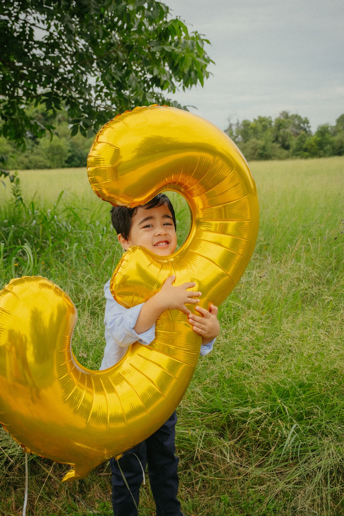 Large golden number three balloon photographed in grassy field with lush green trees in the background.