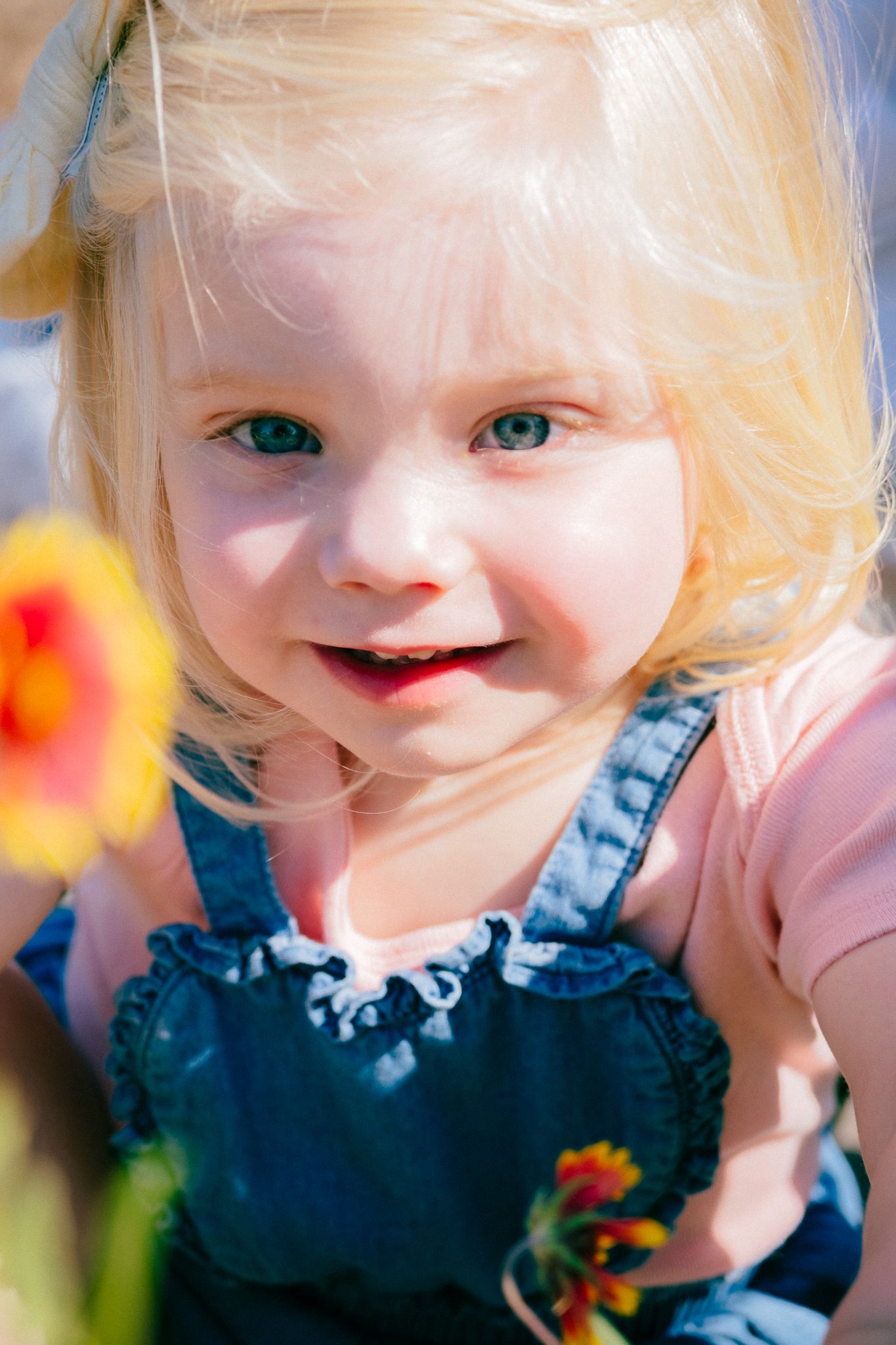 A young child with blonde hair and blue eyes smiles at the camera while flowers bloom in soft focus behind them.
