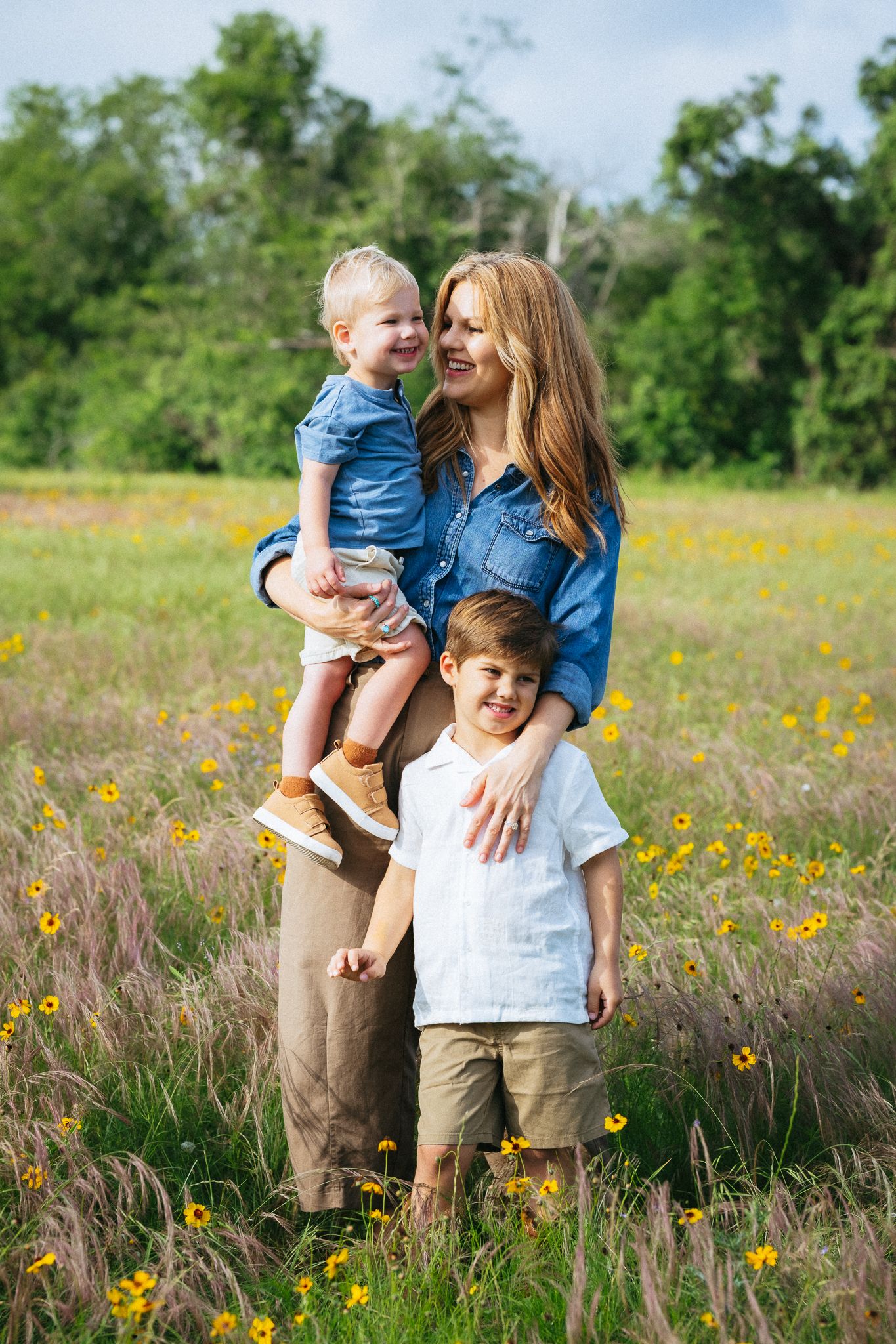 A mother holds a toddler while her young son stands beside her in a sunny wildflower meadow.