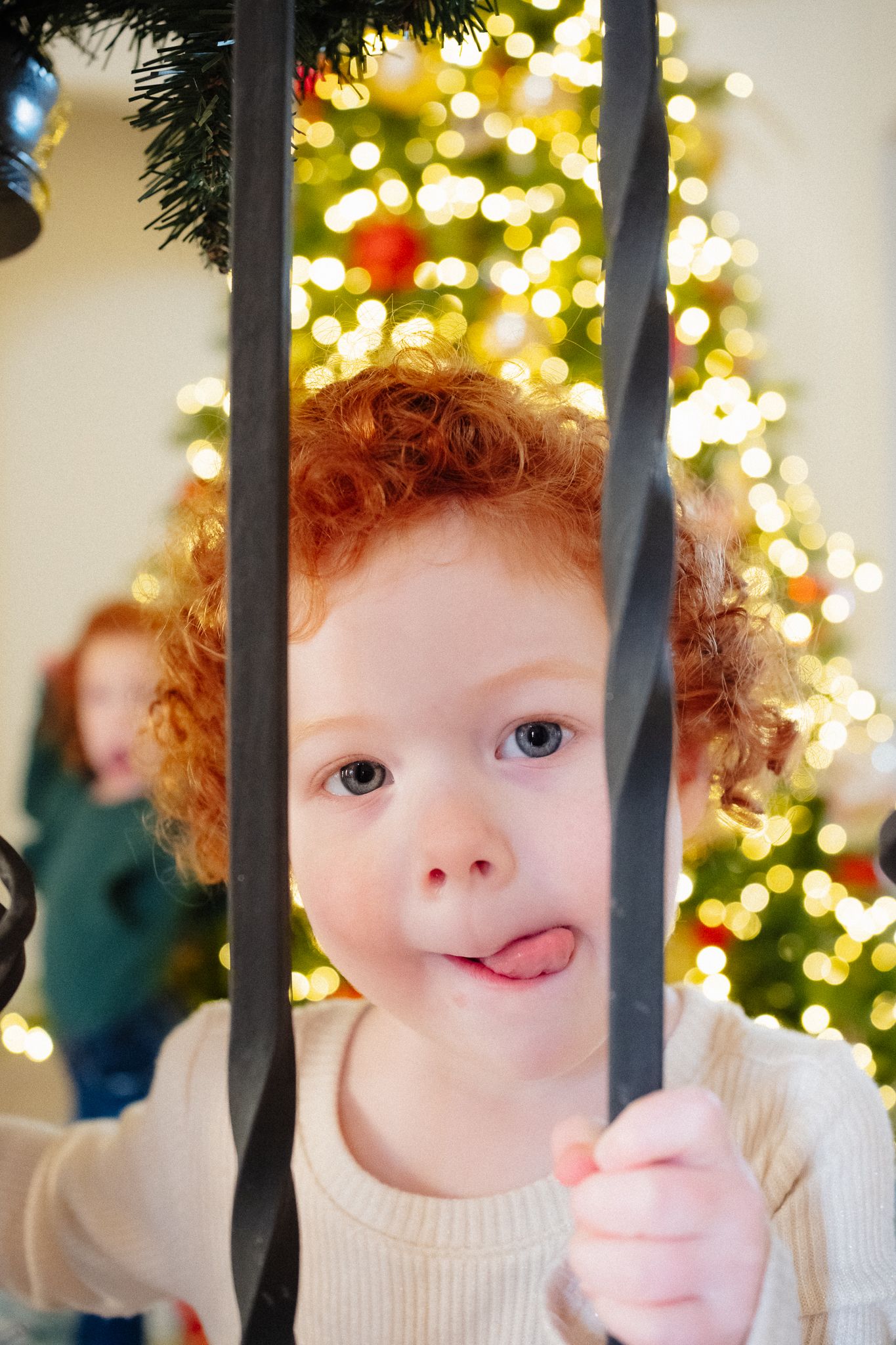 Curly red-haired child sticking out tongue behind black railings with Christmas tree lights in background.