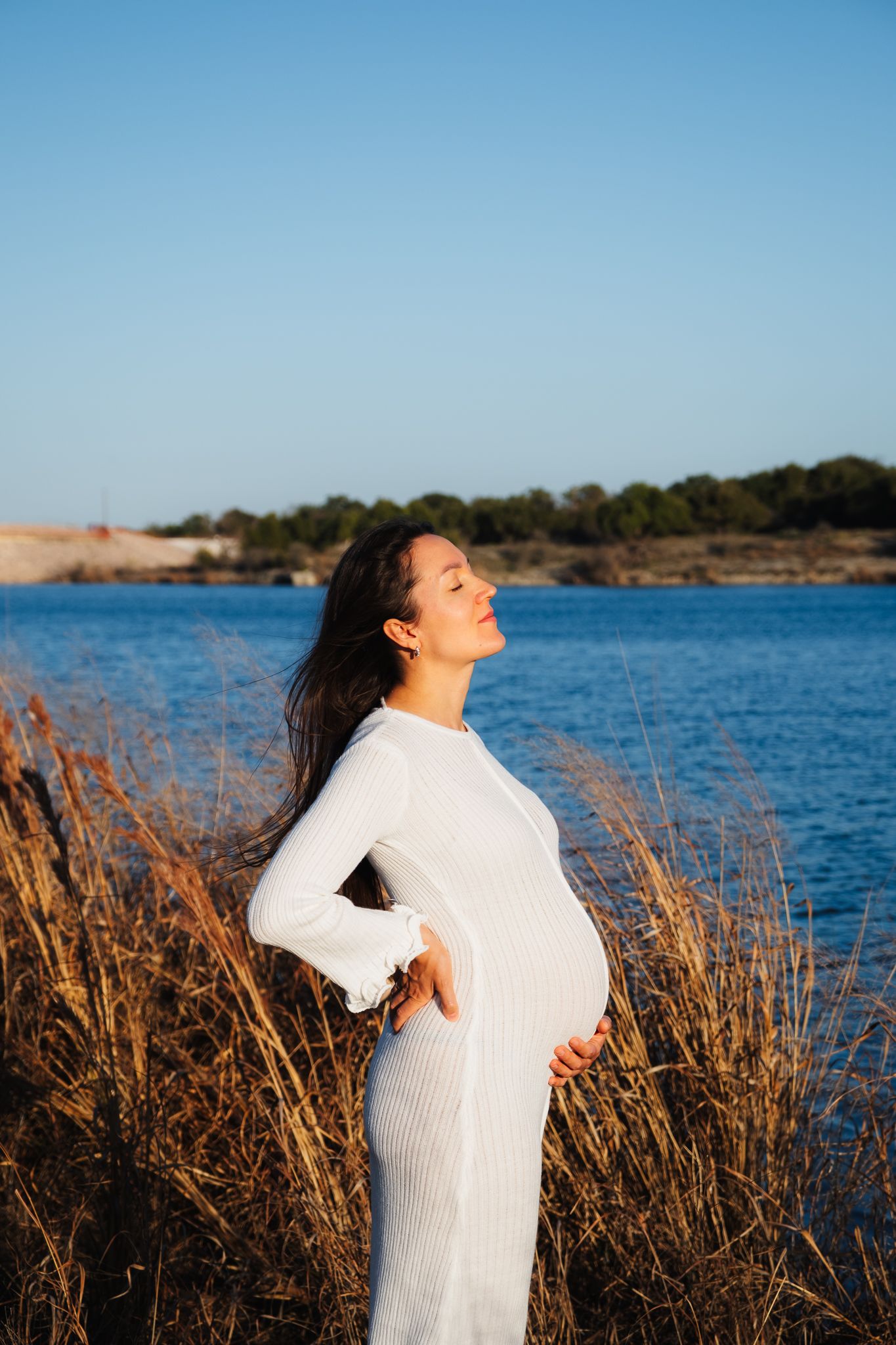 A pregnant woman in a white dress poses by a lake at sunset among tall grass and water reeds.