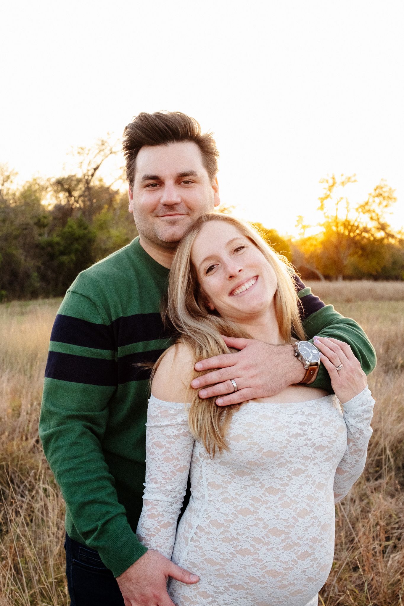 A couple embraces in a field during golden hour sunset, wearing a green sweater and white lace top.