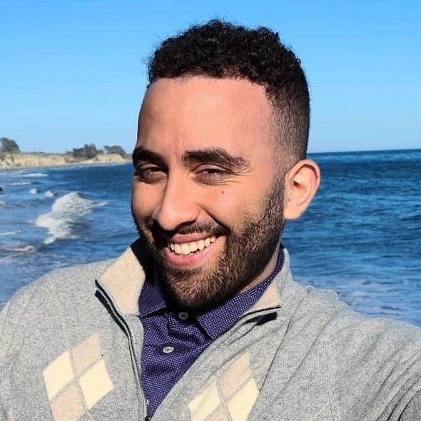 Person smiling at the beach with ocean waves and coastline visible in the background.