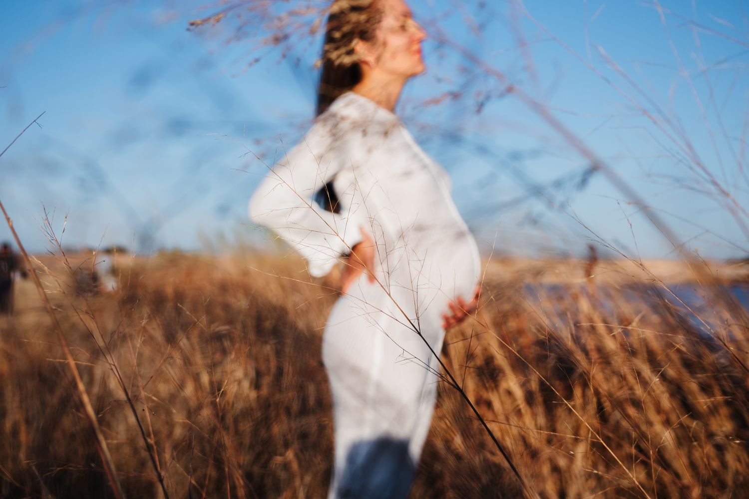 A figure in a flowing white dress stands among tall prairie grass against a bright blue sky, creating a dreamy silhouette.